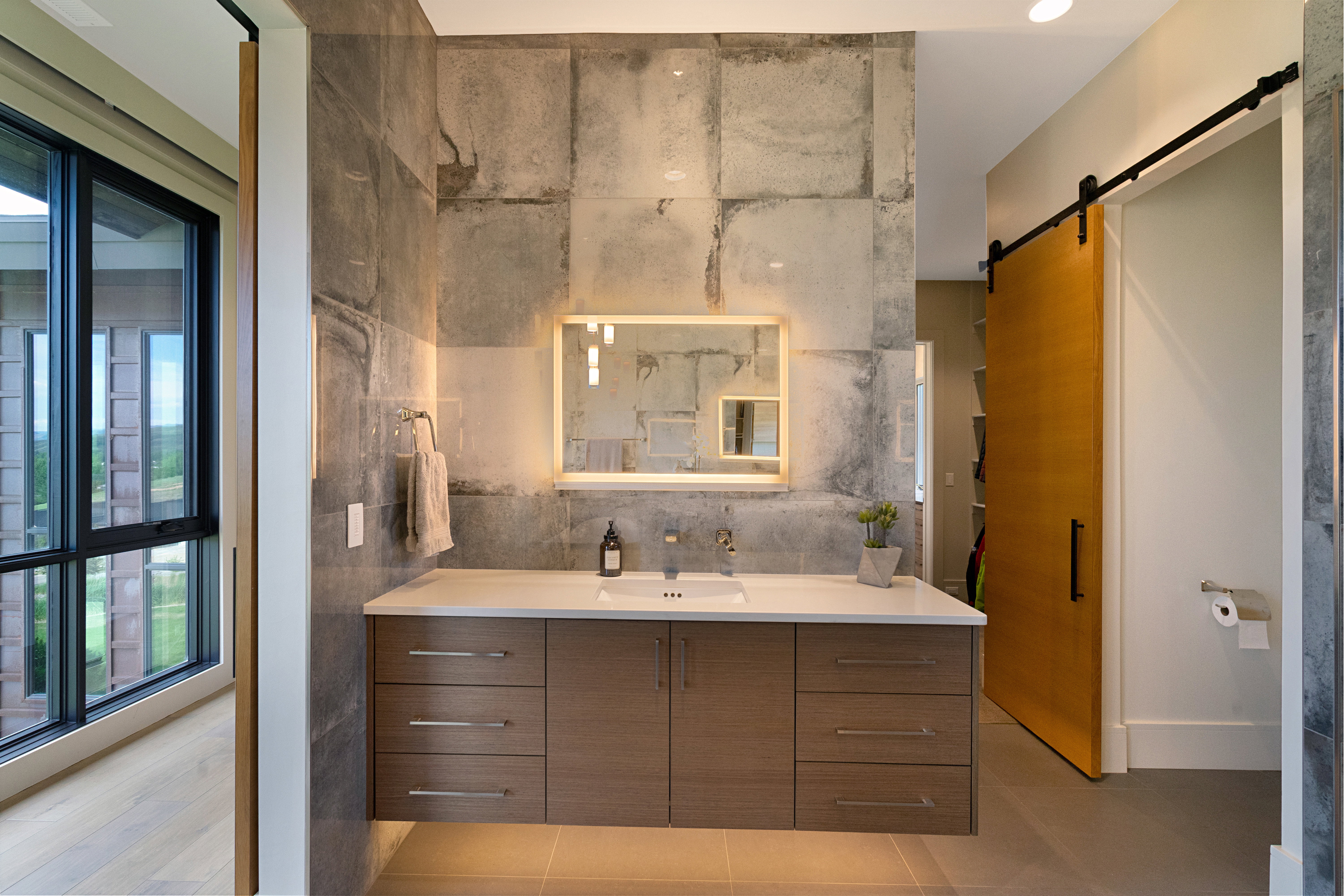 Modern bathroom vanity featuring a light-toned countertop with an undermount sink, contemporary wood-finish cabinetry, a backlit mirror, and a tile-accent wall