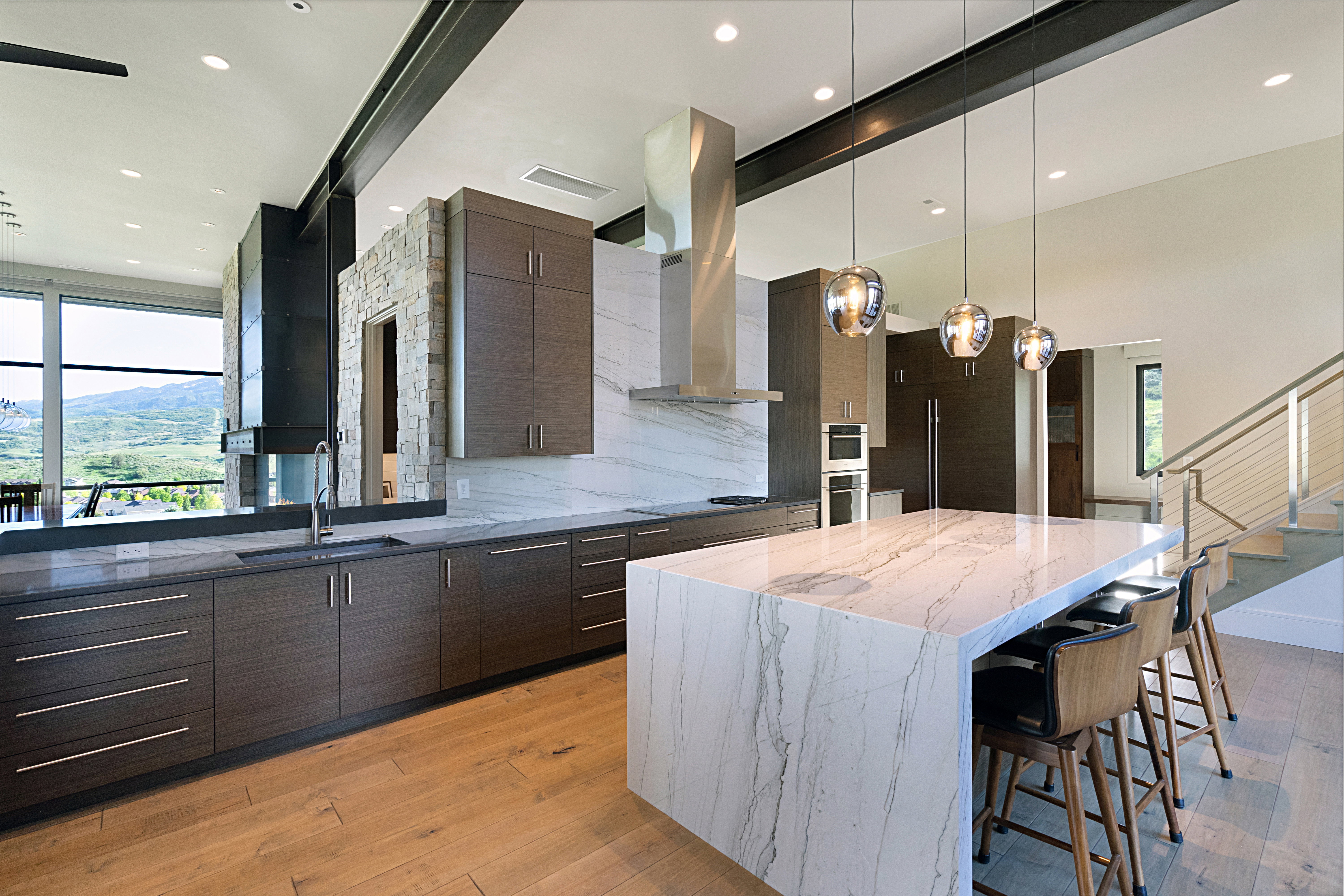 Spacious kitchen featuring an oversized island with waterfall countertop, wood-finish flooring, dark wood-finish cabinetry, stainless steel appliances, and a full-height stone backsplash