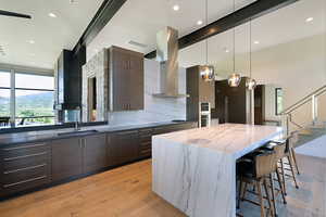 Spacious kitchen featuring an oversized island with waterfall countertop, wood-finish flooring, dark wood-finish cabinetry, stainless steel appliances, and a full-height stone backsplash