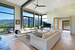 Spacious living area featuring floor-to-ceiling windows, high ceilings with exposed structural beams, light wood-finish flooring, recessed lighting, and a modern ceiling fan