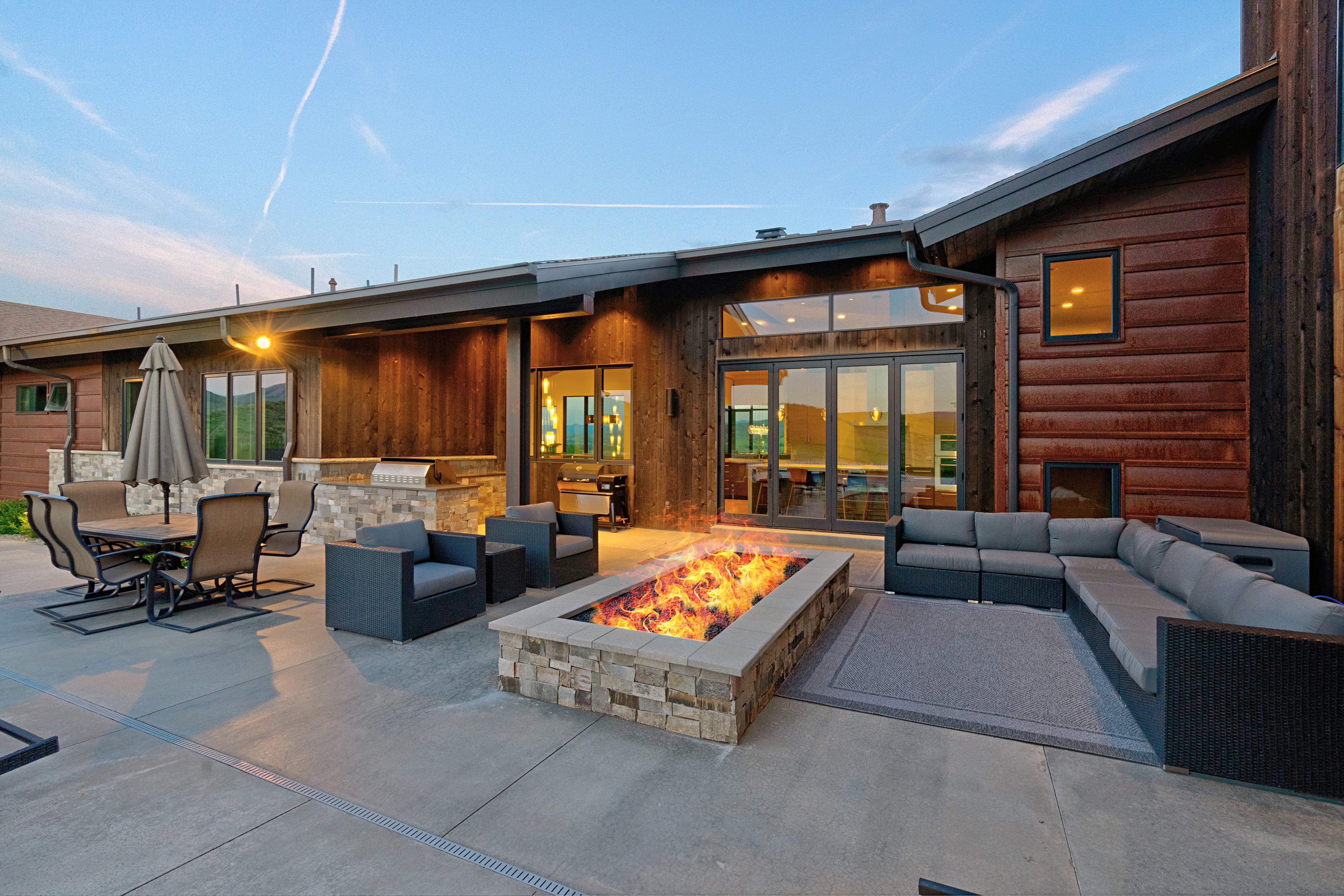 Expansive concrete patio featuring a stone-clad fire pit, built-in outdoor kitchen with a grill, and an umbrella-shaded dining area