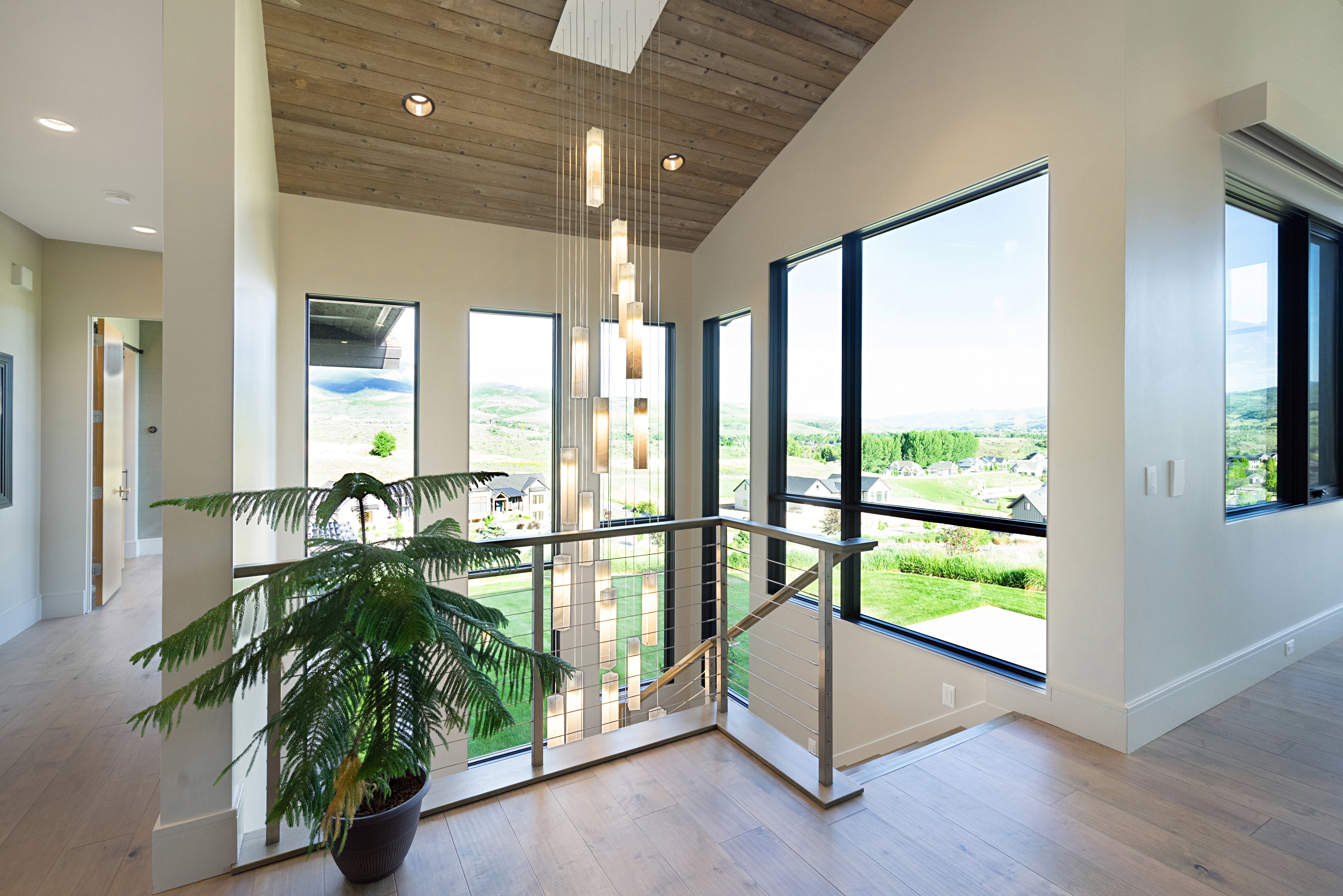 Vaulted wood-finish ceiling above an open landing featuring a multi-tiered chandelier, extensive window placement, stainless steel and cable railing, and wood-finish flooring