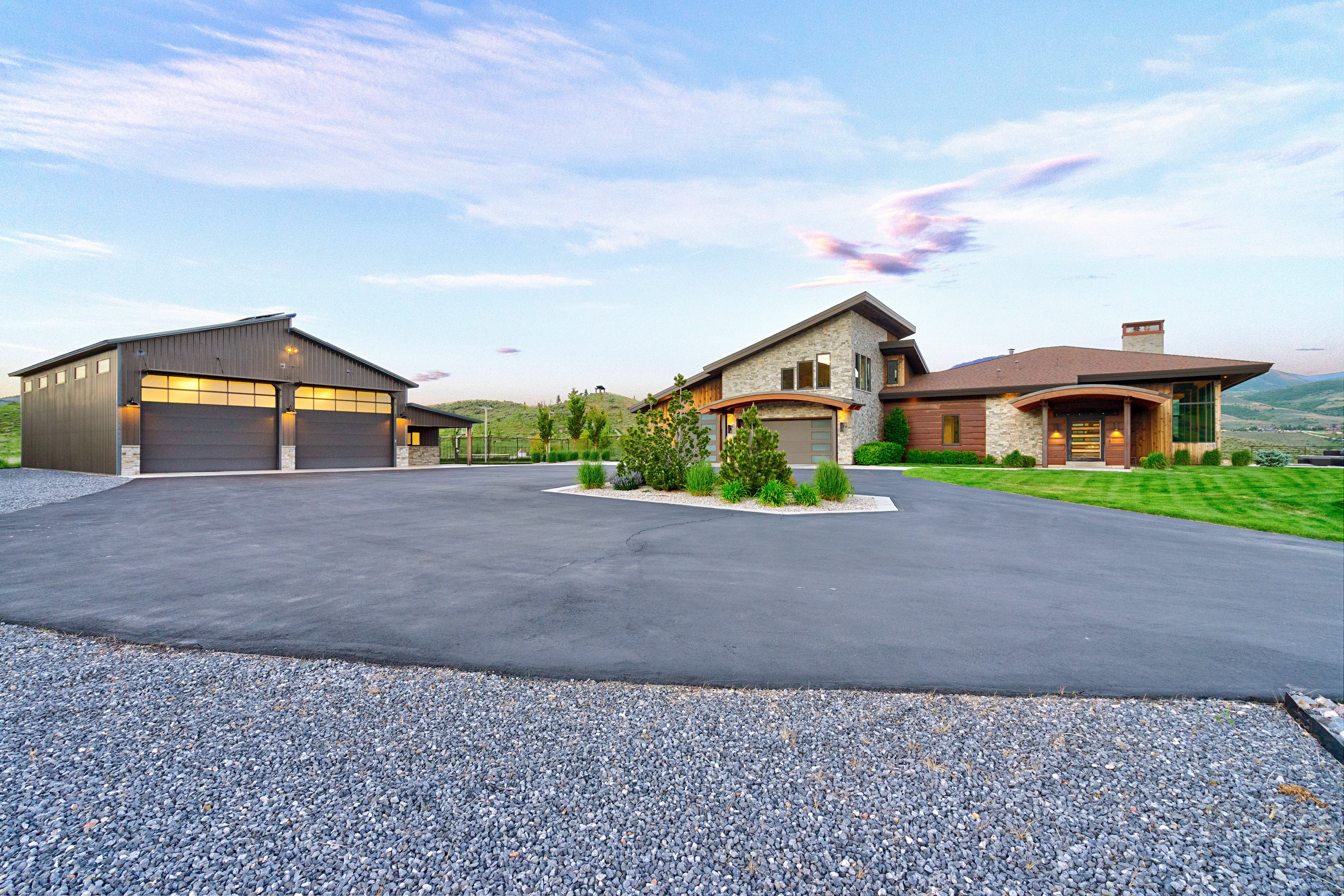 Modern architectural residence with an expansive driveway, a separate multi-car garage, stone and wood-panel exterior accents, and a landscaped property