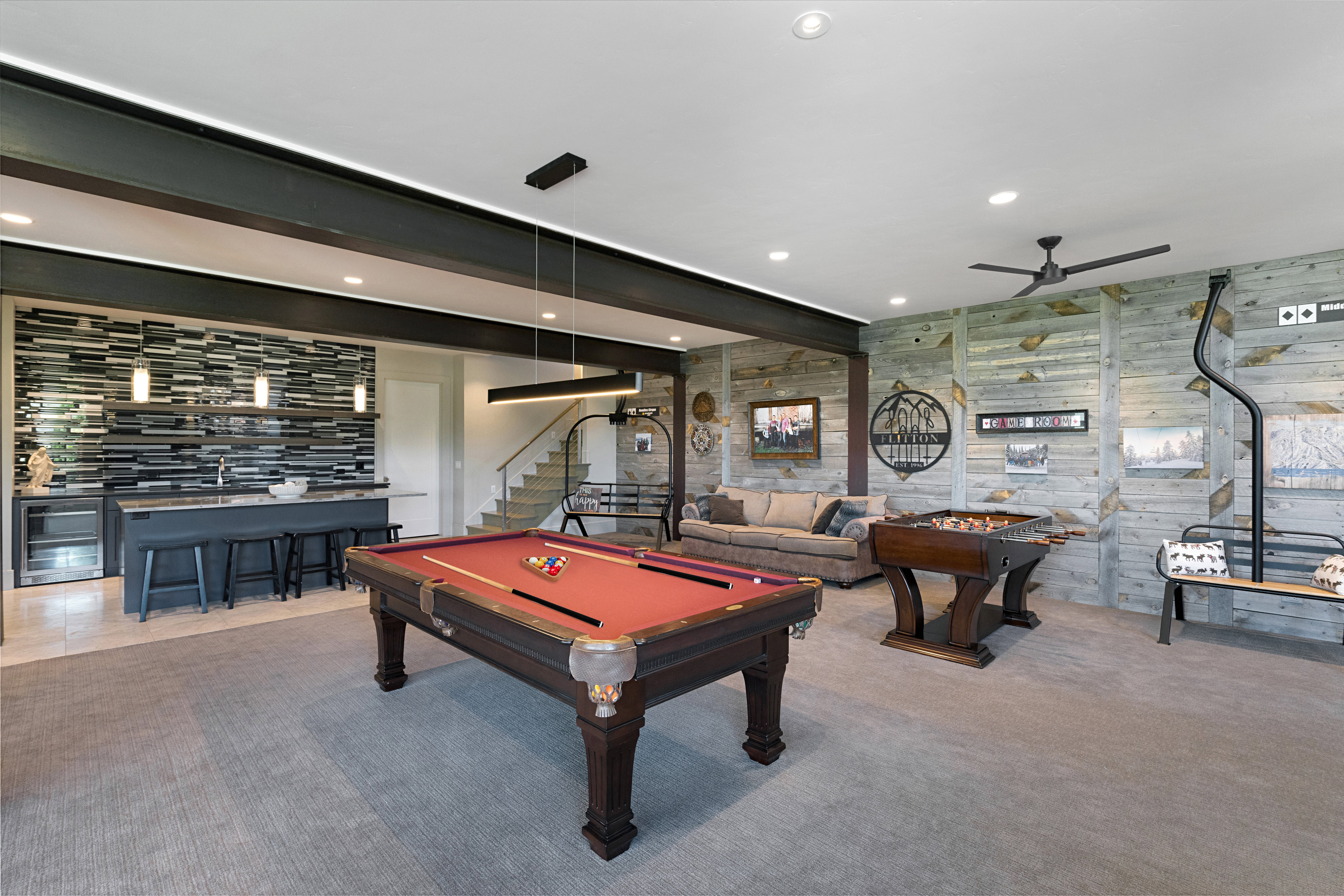 Spacious recreation area featuring a wet bar with a tiled accent wall and built-in cabinetry, exposed structural beams, recessed lighting, and wood-finish wall paneling