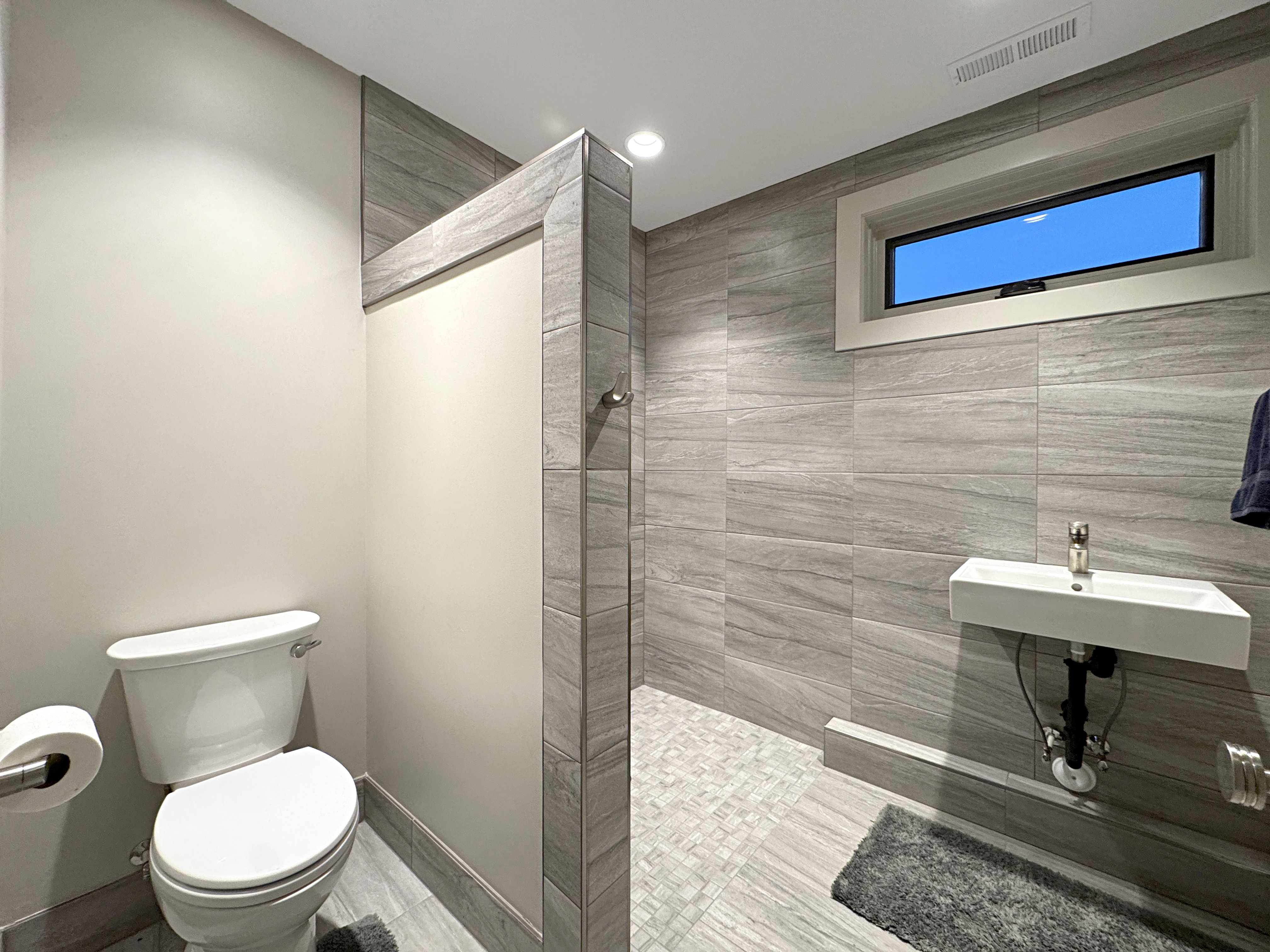 Bathroom featuring a wall-mounted rectangular sink, large format tile walls, a toilet, a privacy partition, and an elevated rectangular window