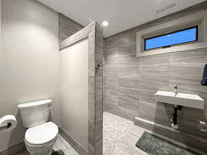 Bathroom featuring a wall-mounted rectangular sink, large format tile walls, a toilet, a privacy partition, and an elevated rectangular window