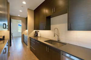 Galley kitchen featuring dark wood-finish cabinetry, light wood-finish flooring, undermount stainless steel sink with gooseneck faucet, light solid surface countertops, and white subway tile backsplash