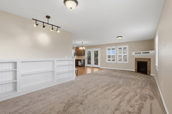 Carpeted living space featuring a fireplace with a stone surround, built-in shelving, two windows with shutters, and a track lighting fixture