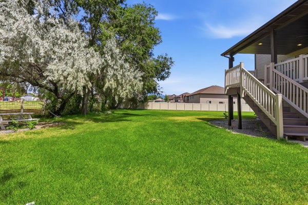 Expansive green lawn featuring mature trees, a multi-level deck with stairs, a neutral-toned privacy fence, and a partially visible picnic table
