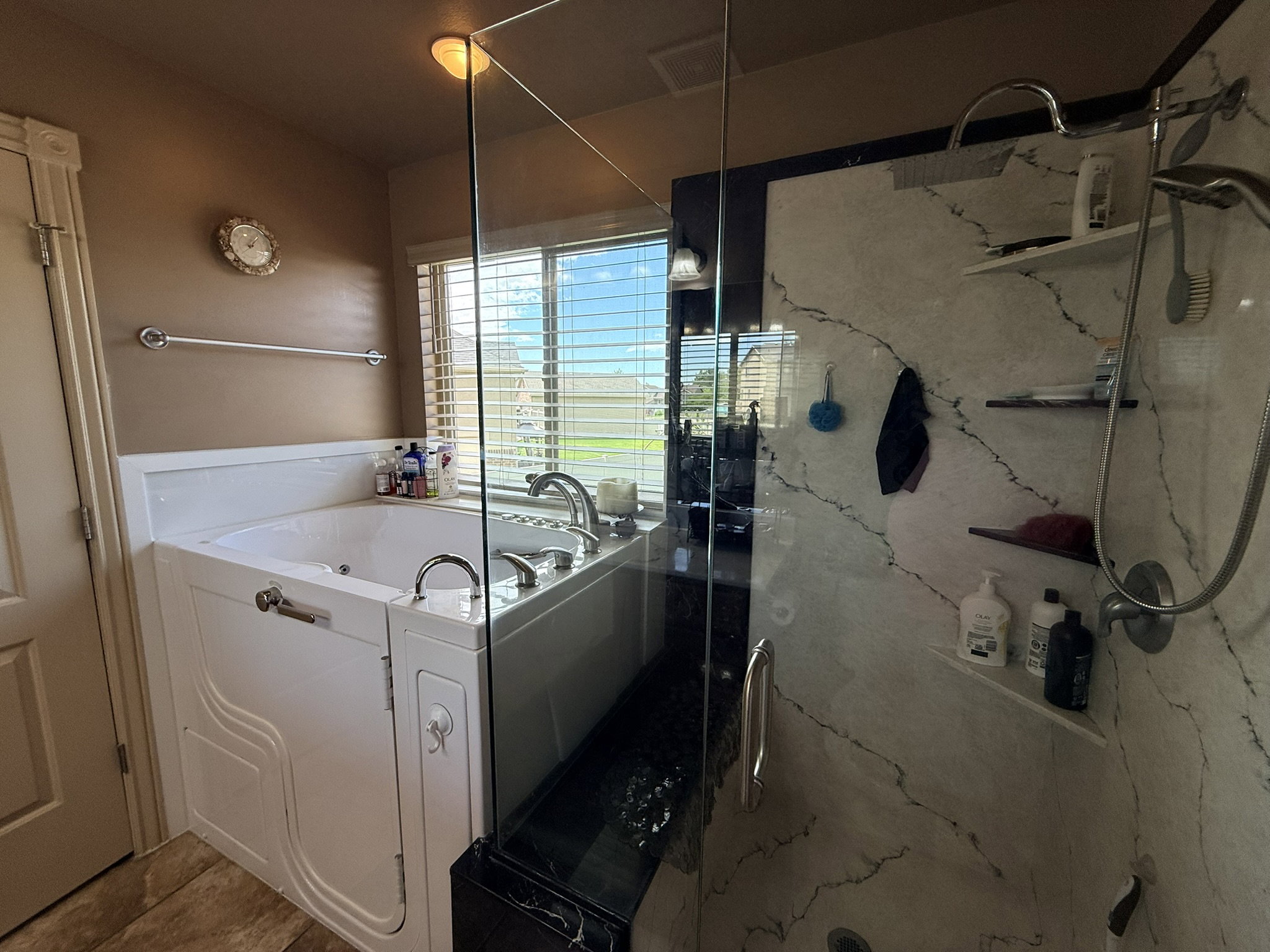 Walk-in tub with chrome fixtures, accompanied by a glass-enclosed shower featuring white and gray veined wall panels, a rainfall showerhead, and integrated shelving