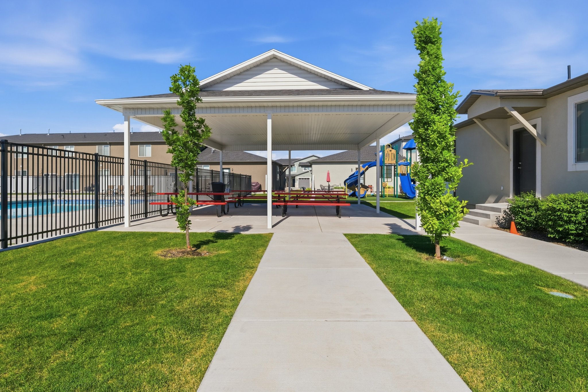 Covered outdoor recreation area featuring a paved walkway, green lawn, a playground, and a fenced swimming pool