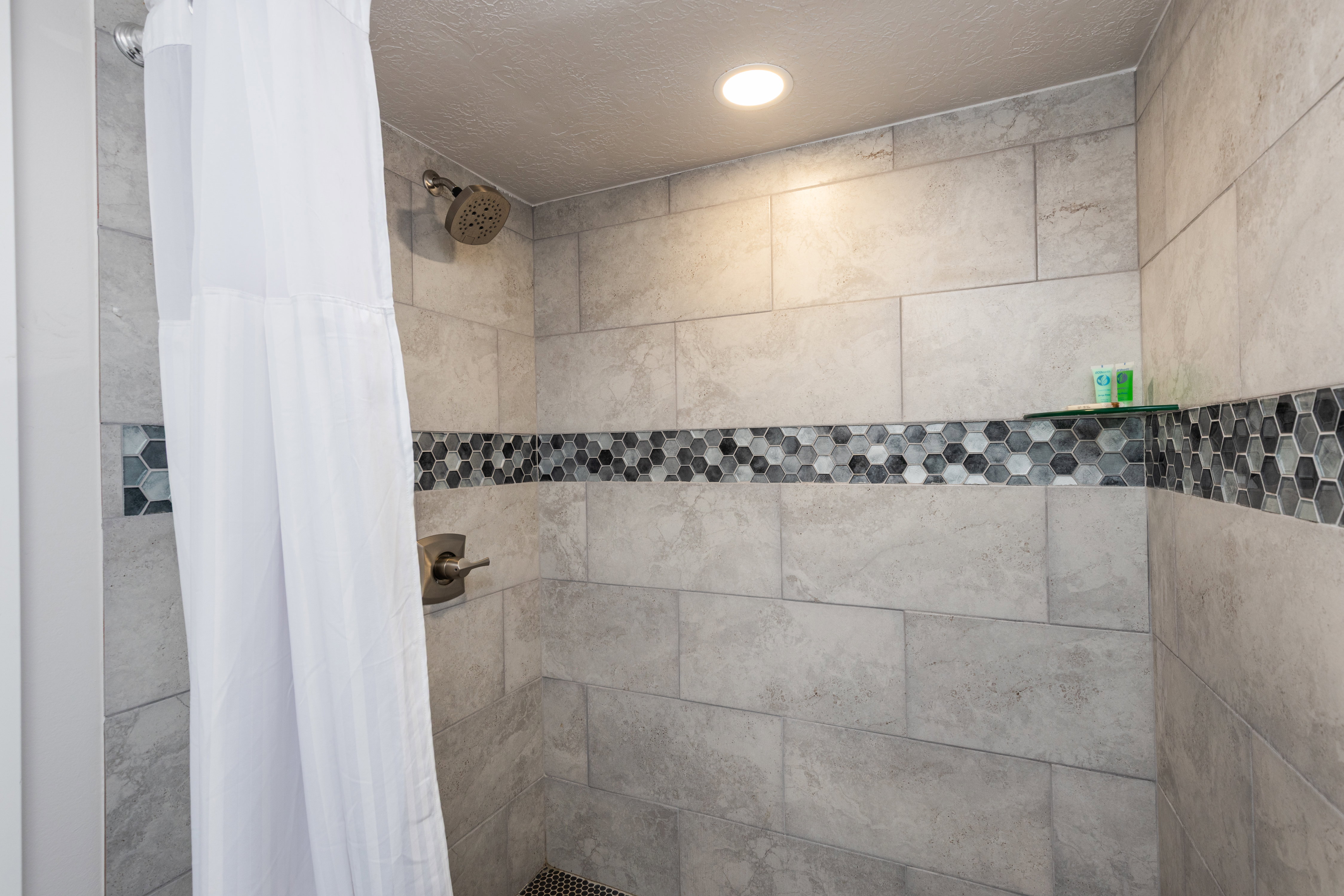 Walk-in shower featuring large format tile walls, a decorative hexagonal mosaic tile band, a recessed niche with hexagonal tile, a glass shelf, and a ceiling-mounted recessed light