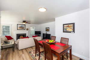 Open-concept living and dining area featuring wood-finish flooring, a ceiling fan, a fireplace with a white mantel and dark stone surround, and multiple windows with blinds
