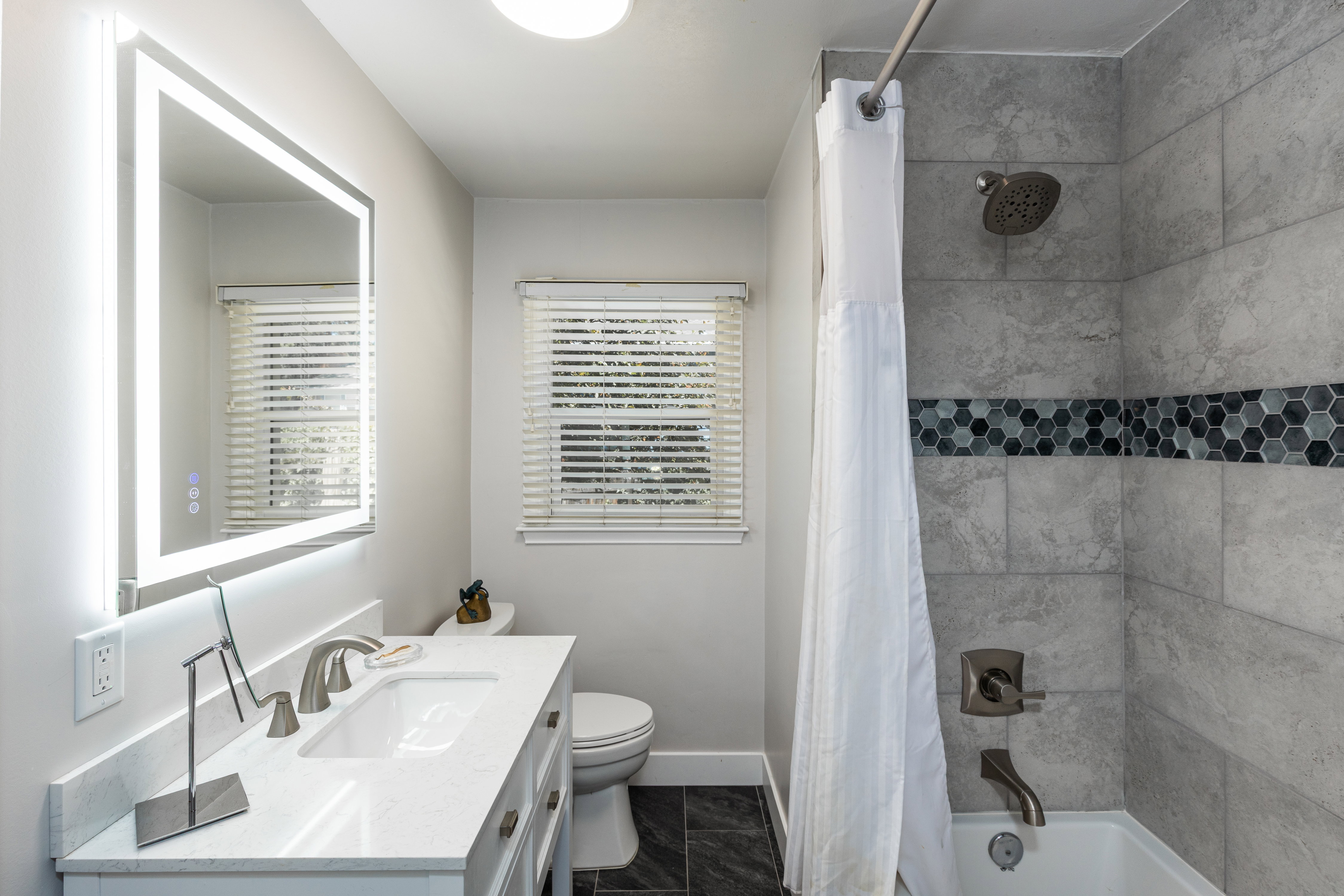 Modern bathroom featuring a back-lit vanity mirror, white countertop, and a built-in tub with a tiled shower surround