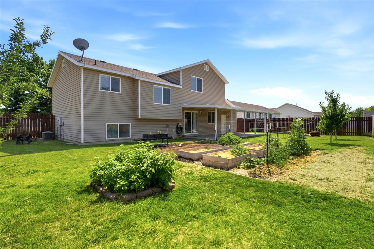 Expansive fenced backyard featuring a covered patio, multiple raised garden beds, a mature tree, and a lush lawn