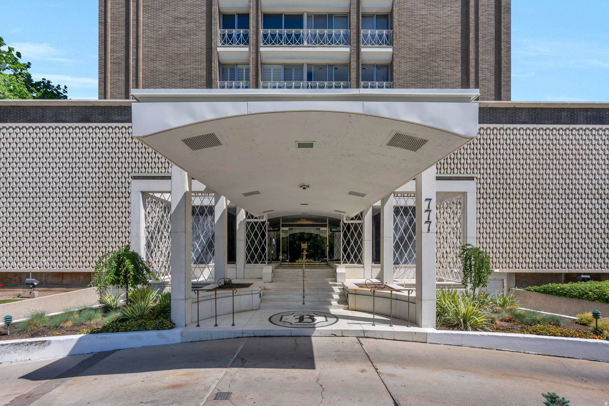 Building entrance featuring a curved portico, patterned facade, integrated planters with landscaping, and a concrete driveway