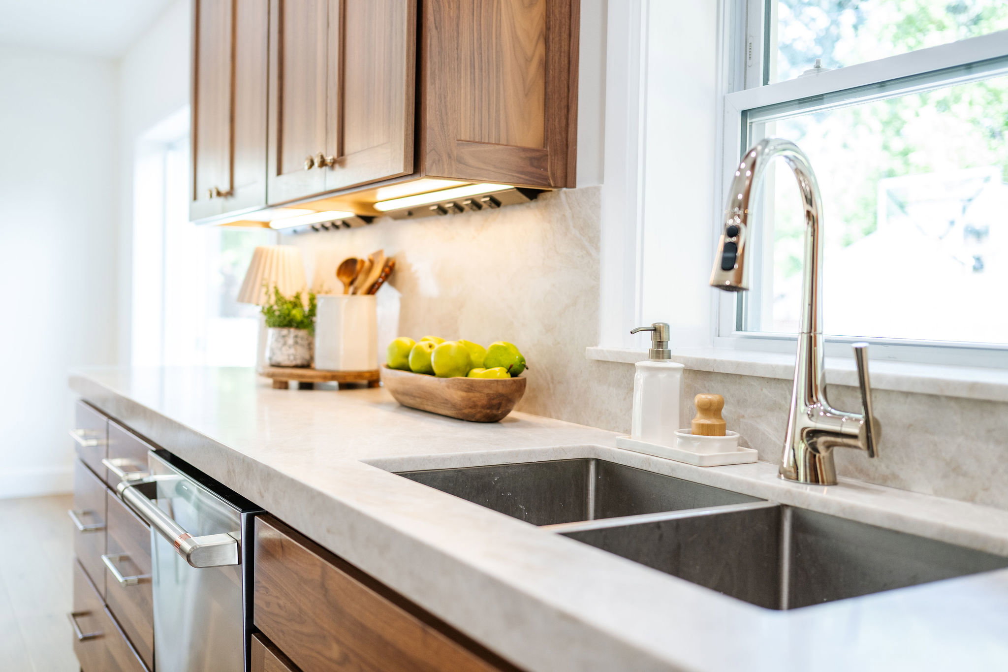Kitchen featuring a double basin stainless steel sink, a chrome gooseneck faucet, light-toned stone countertops, natural wood-finish cabinetry, and under-cabinet lighting