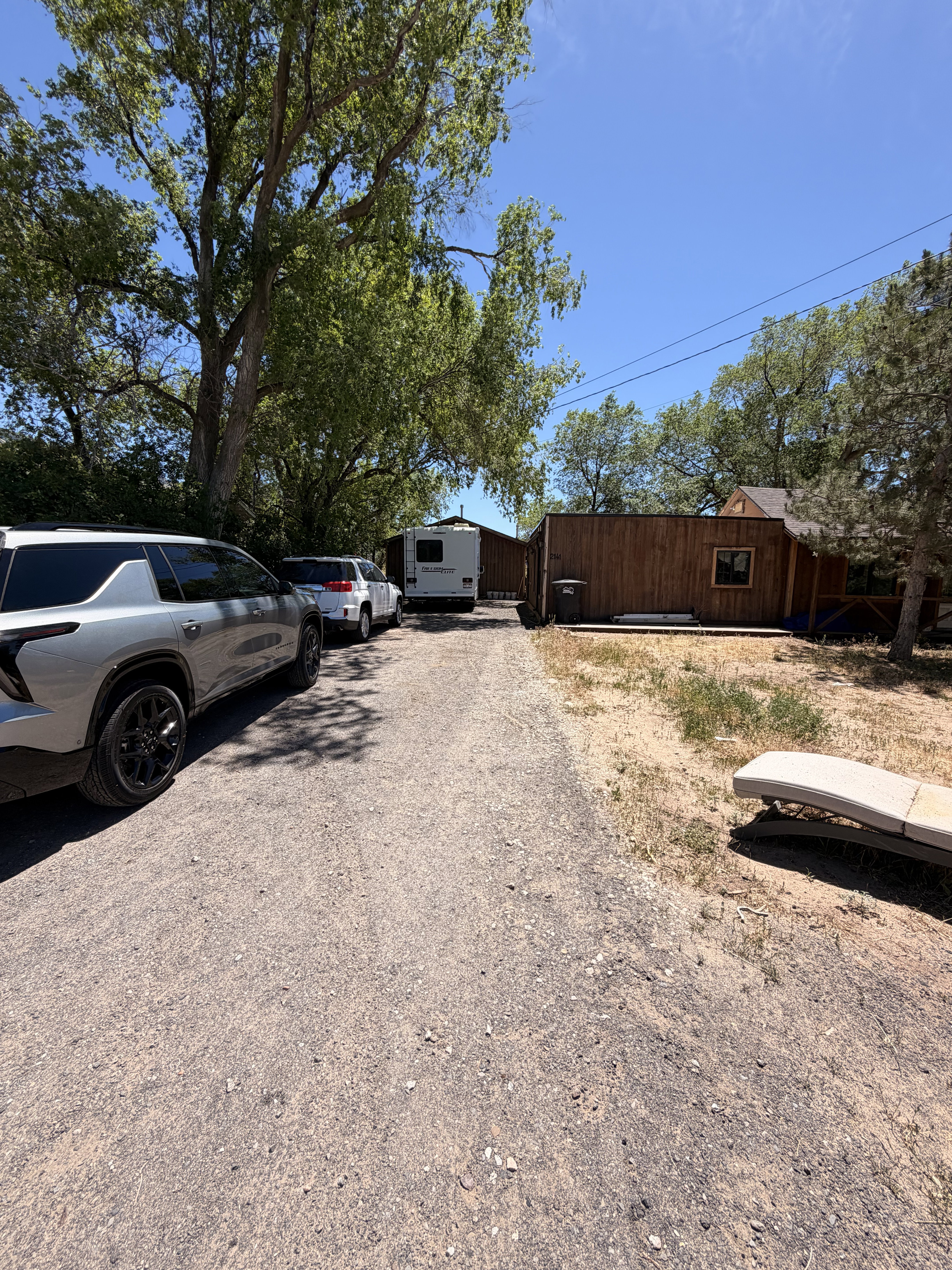 Gravel driveway offering ample parking space, bordered by mature trees and a wood-sided structure