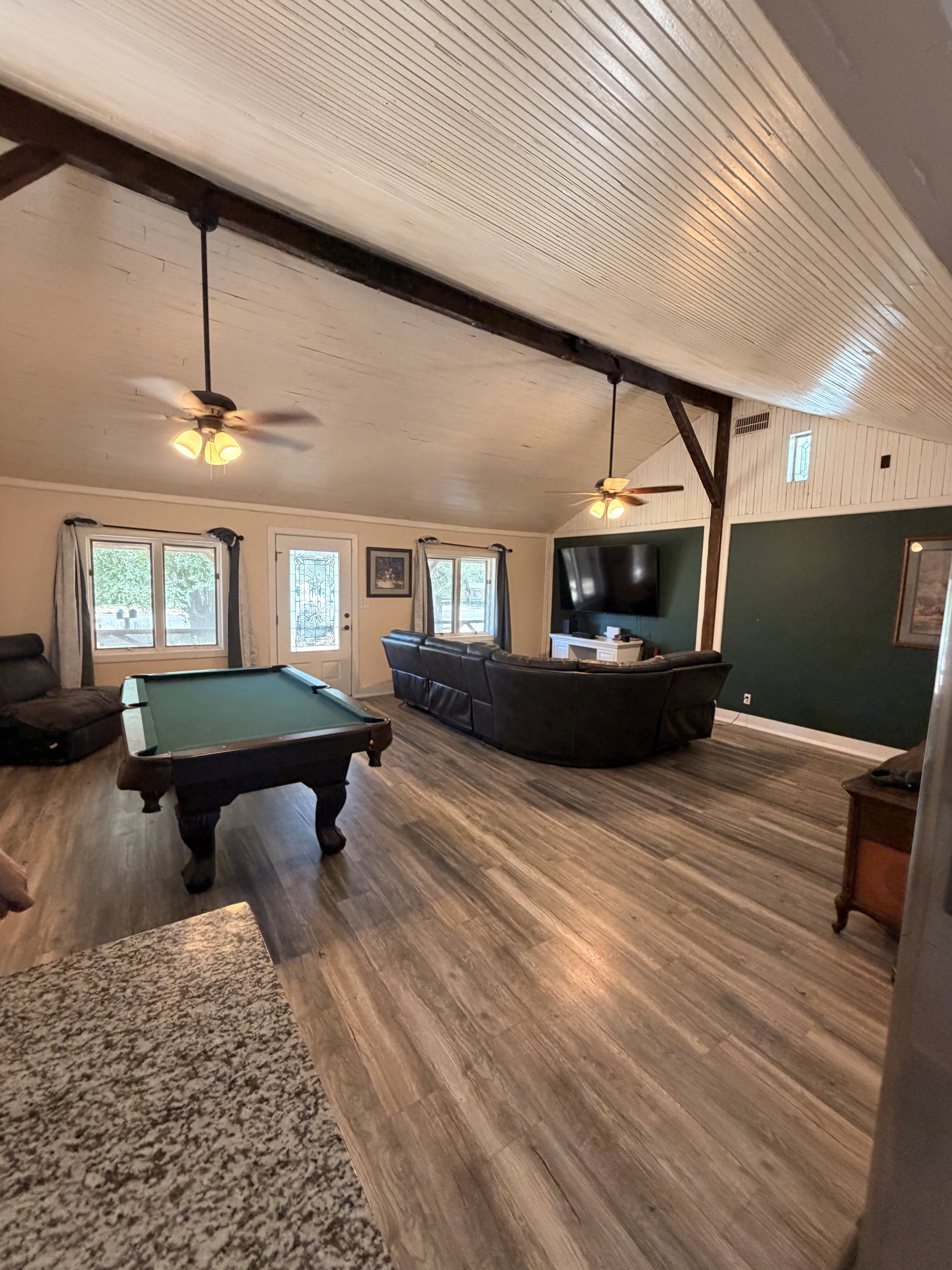 Spacious open-concept room featuring vaulted ceilings with exposed beams, wood-finish flooring, and multiple windows