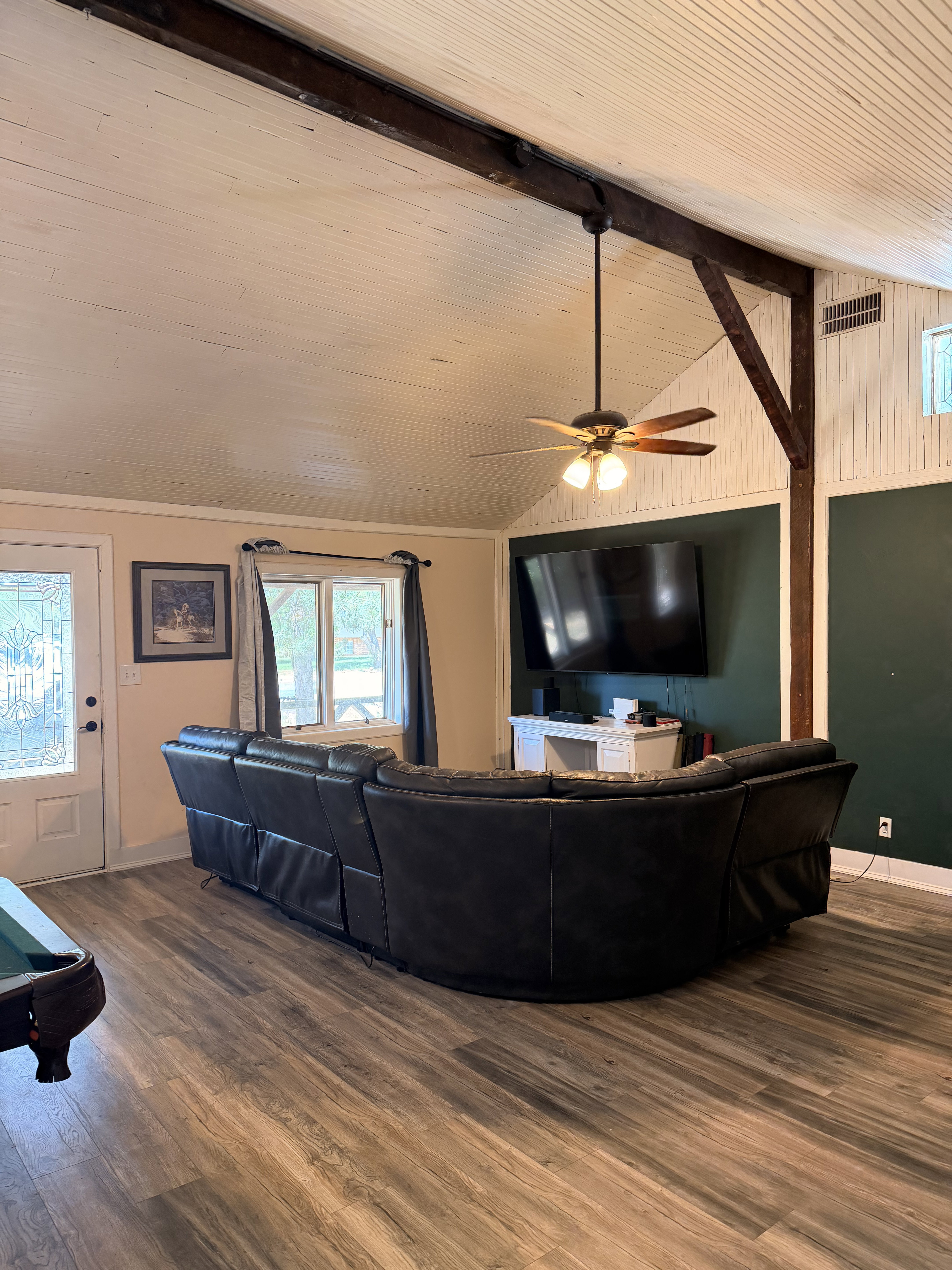 Spacious living area featuring wood-finish flooring, exposed ceiling beams, a vaulted tongue-and-groove ceiling, and a ceiling fan with integrated lighting
