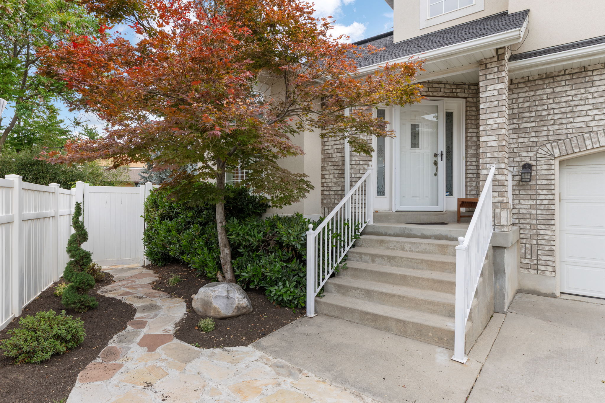 Front exterior featuring a brick facade, white entry door with sidelights, a paved walkway, and mature landscaping.