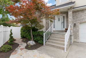 Front exterior featuring a brick facade, white entry door with sidelights, a paved walkway, and mature landscaping.