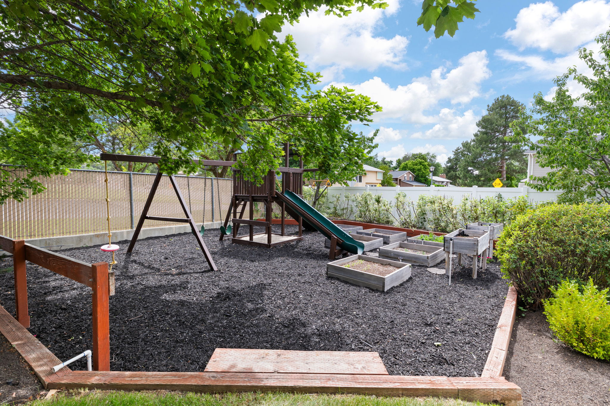 Landscaped backyard featuring a swing set, playset with slide, and multiple raised garden beds