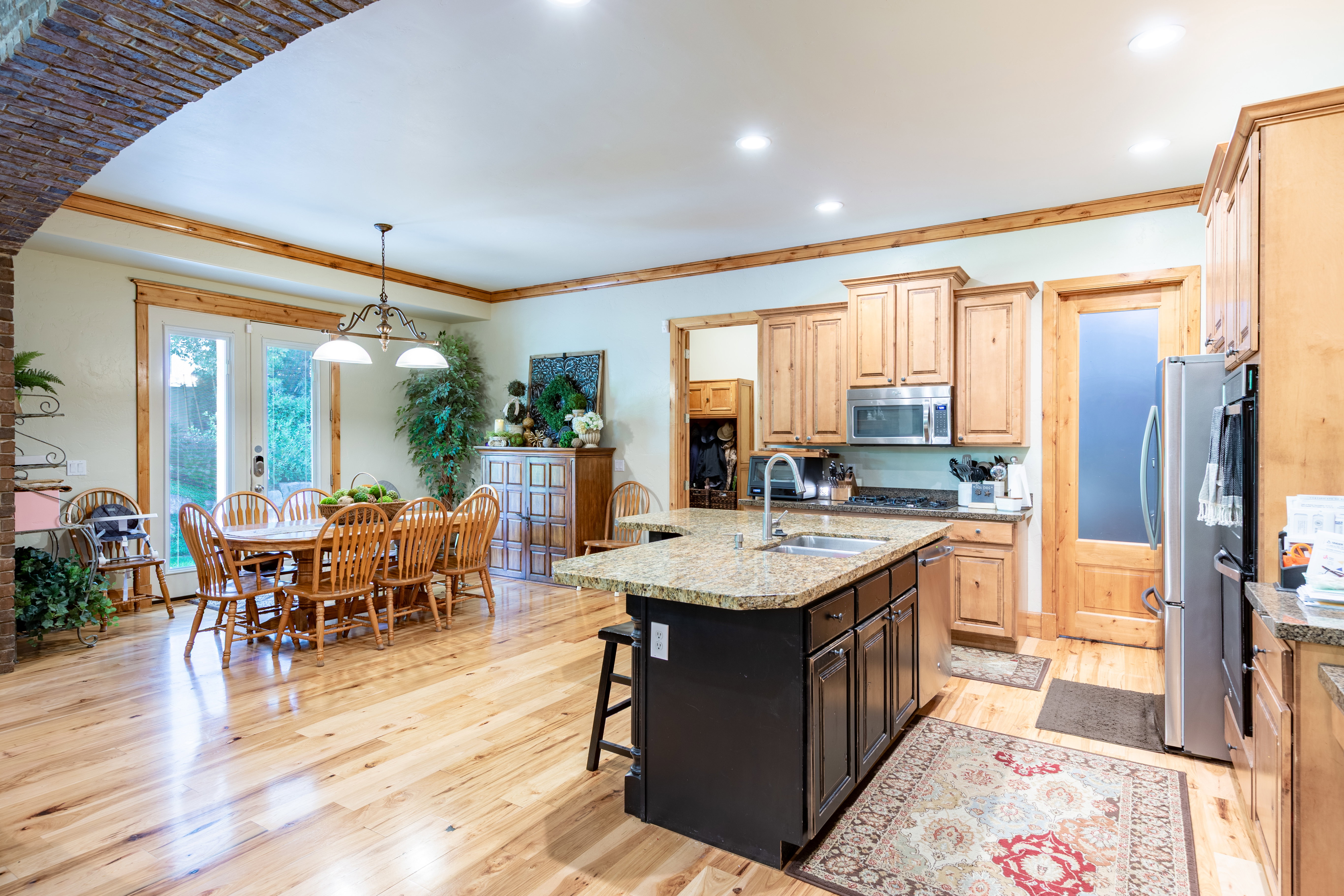 Open-concept kitchen and dining area featuring light wood-finish flooring, a kitchen island with granite-style countertop, natural wood cabinetry, and a brick-finish archway