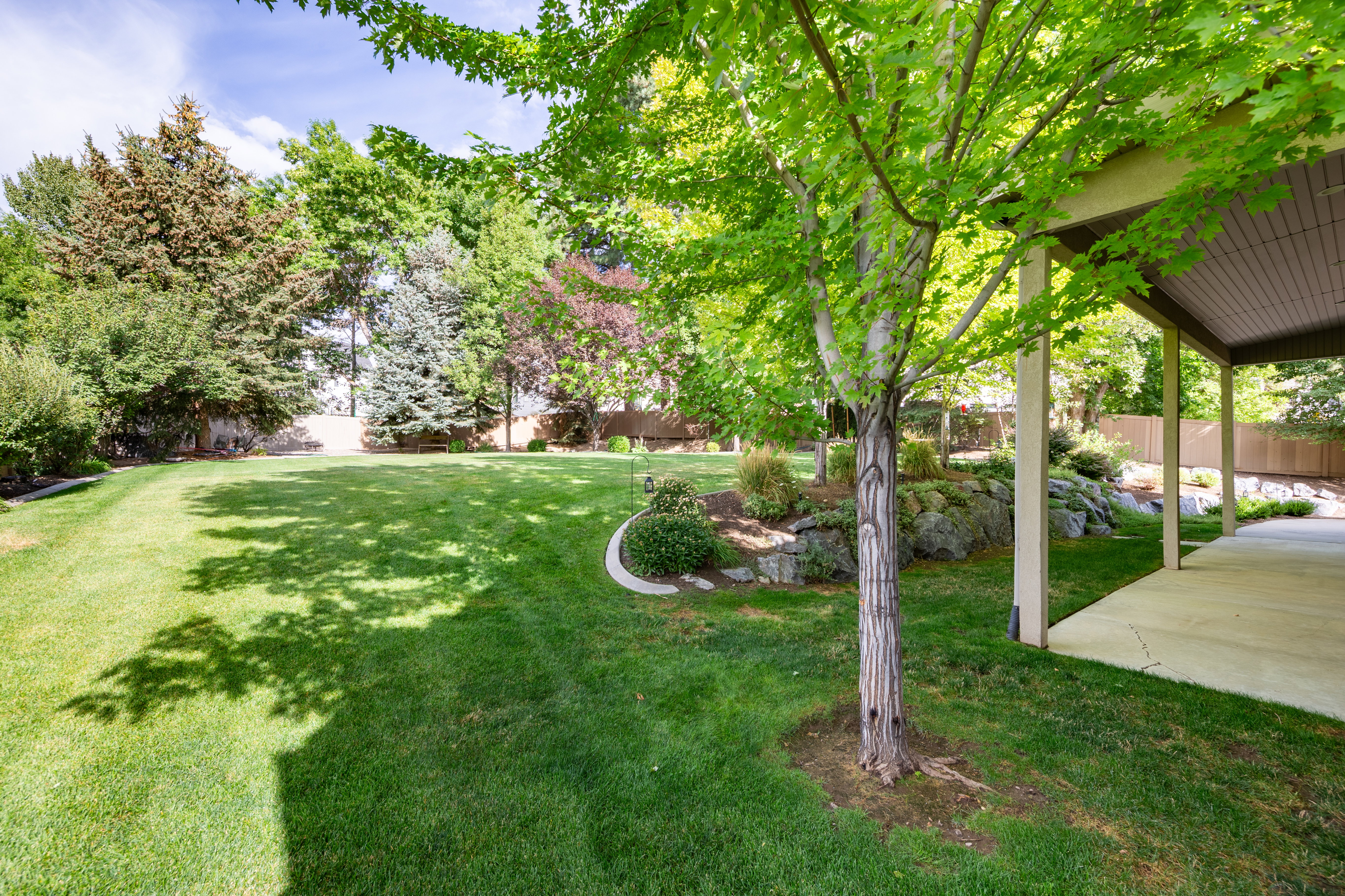 Expansive backyard featuring a lush lawn, mature trees, landscaped rock gardens, and a covered patio