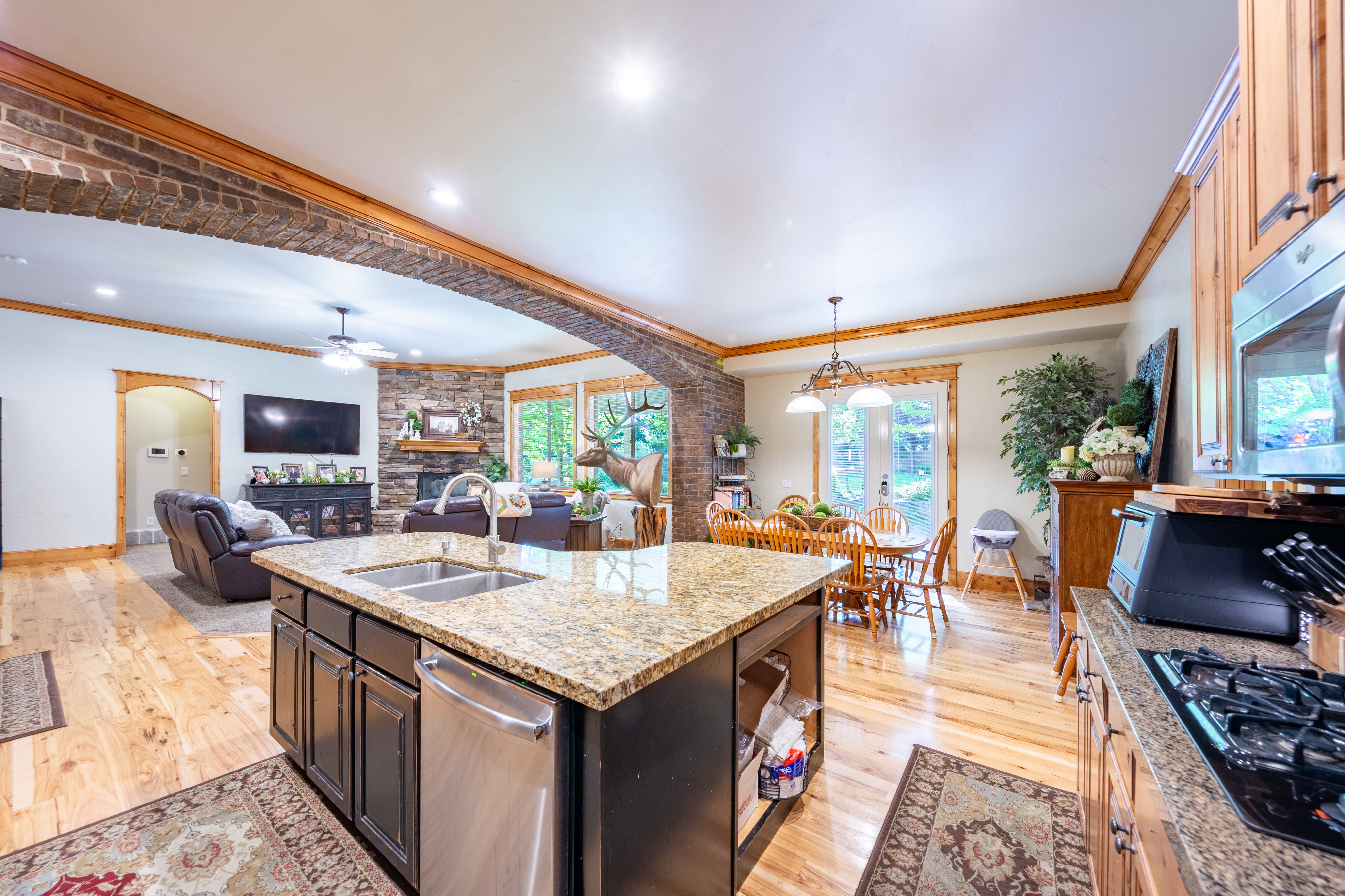Open-concept living space featuring a central kitchen island with a double basin sink and granite-finish countertop, wood-finish flooring, a brick archway, and a stone-clad fireplace