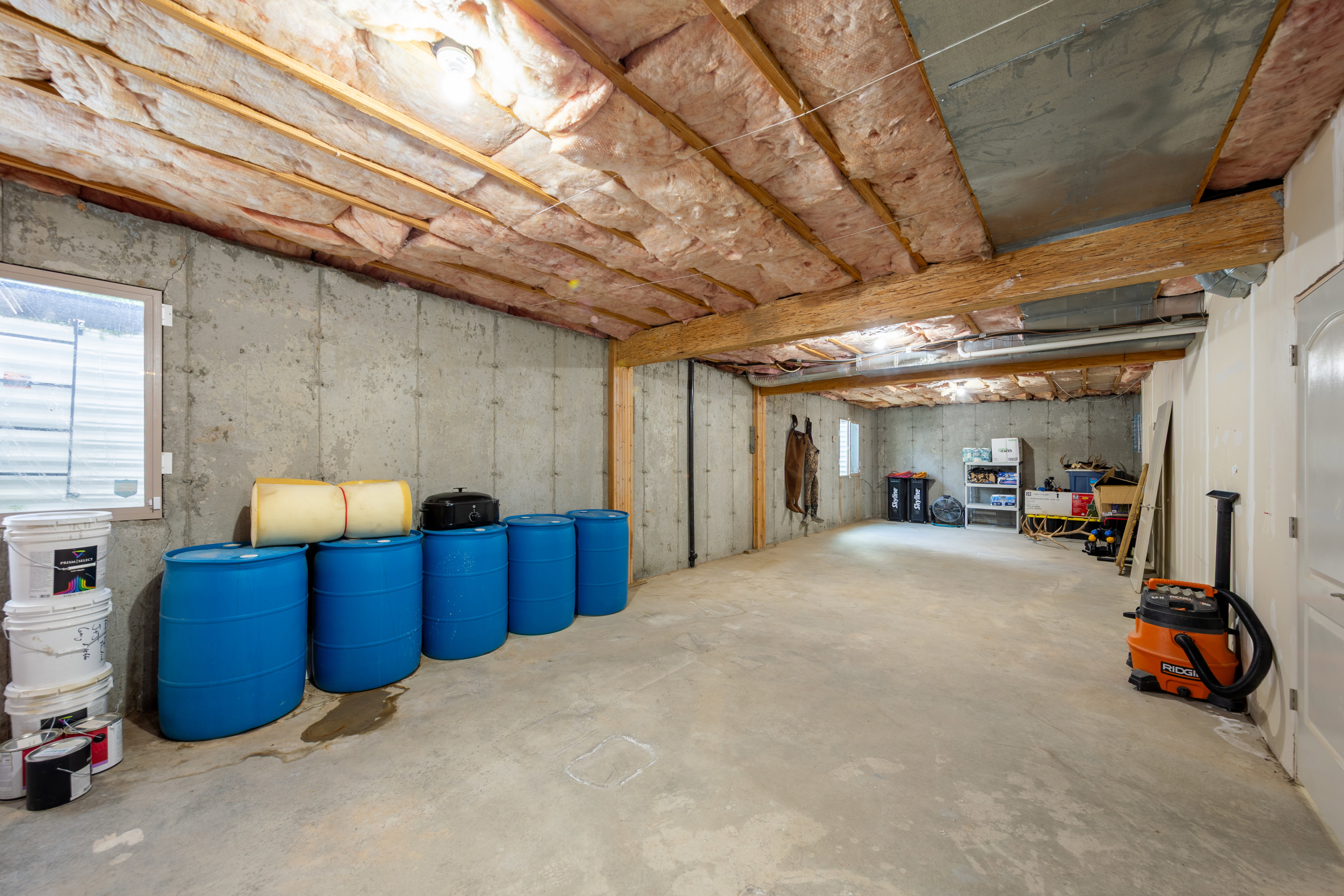 Spacious unfinished basement featuring concrete walls and flooring, exposed wood ceiling joists with insulation, and a visible window for natural light