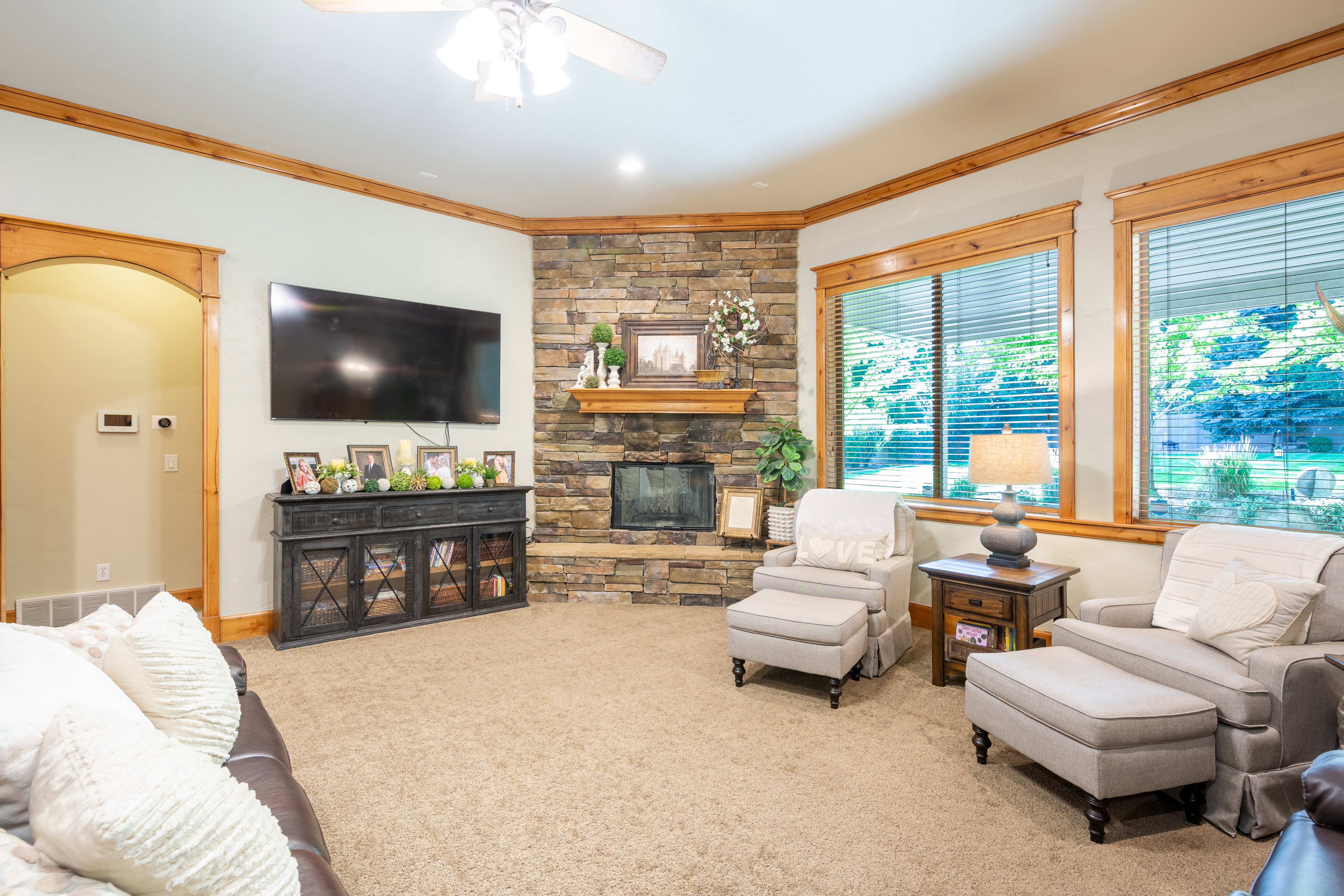 Spacious living area featuring a stone-clad corner fireplace with a wooden mantel, multiple large windows with wooden trim, light tan carpeting, and a ceiling fan