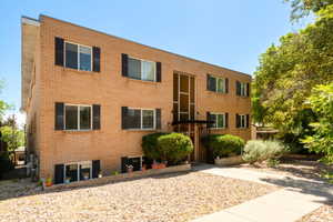 Brick multi-story building featuring multiple windows with dark shutters, a prominent central entrance with an awning, and mature landscaping
