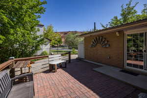 Brick paver patio featuring a sliding glass door entry, a light brick exterior wall, and a raised wooden deck with a cable railing