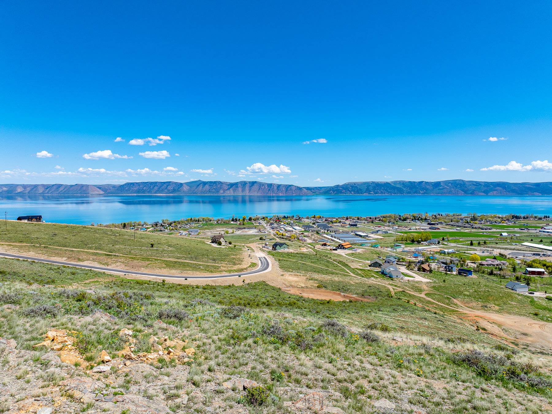Expansive hilltop property featuring a paved road, distant lake, mountainous terrain, and scattered residential structures