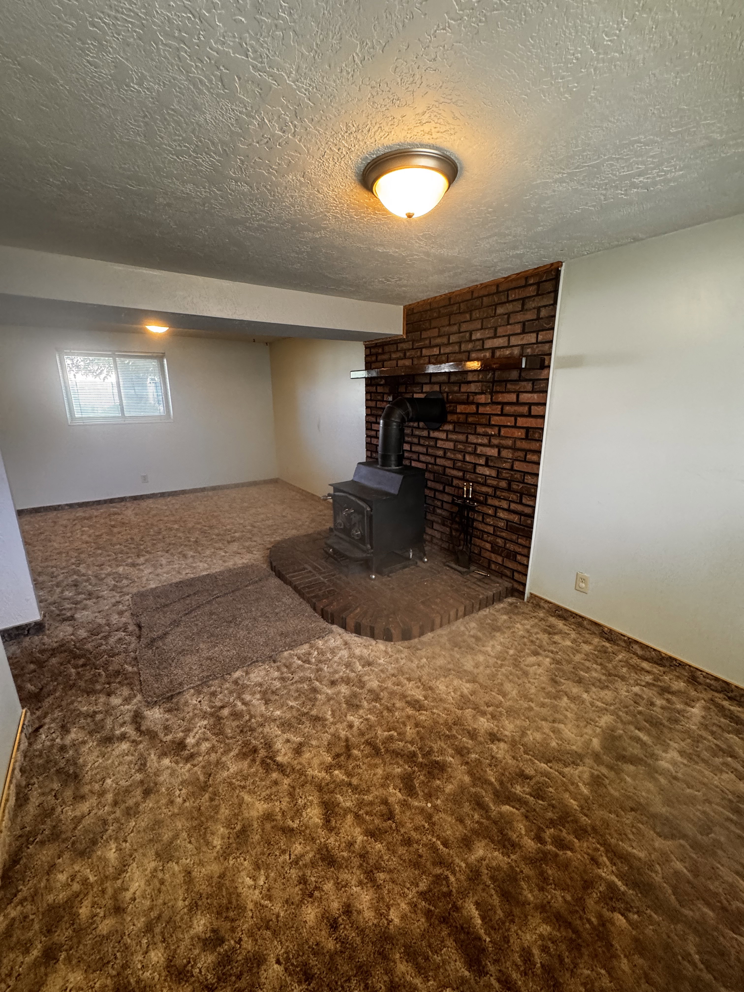 Spacious room featuring a brick accent wall with a wood-burning stove, brown patterned carpet, and a flush mount ceiling light