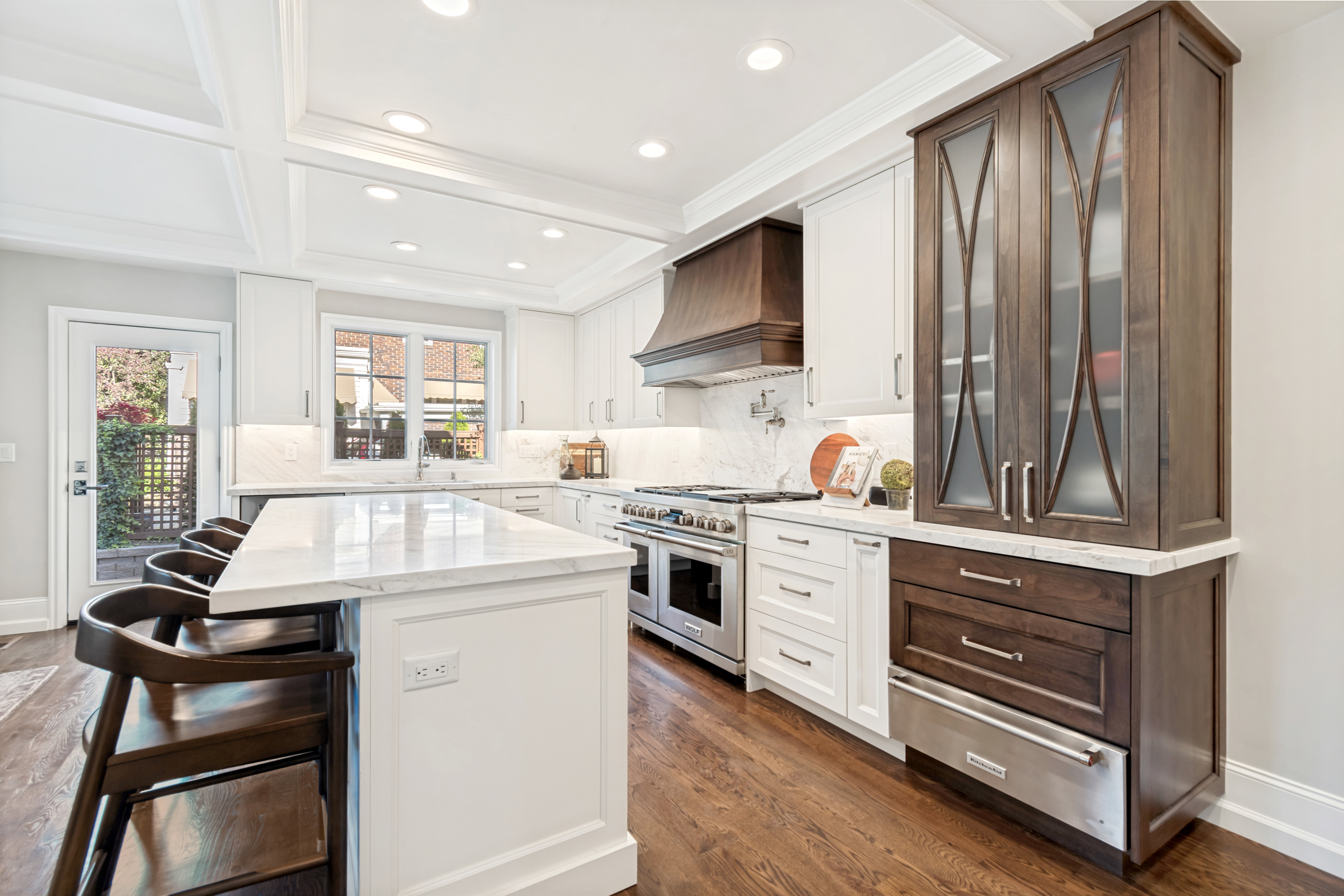 Gourmet kitchen featuring a substantial island with stone countertop, coffered ceiling, stainless steel appliances, custom cabinetry, and wood-finish flooring