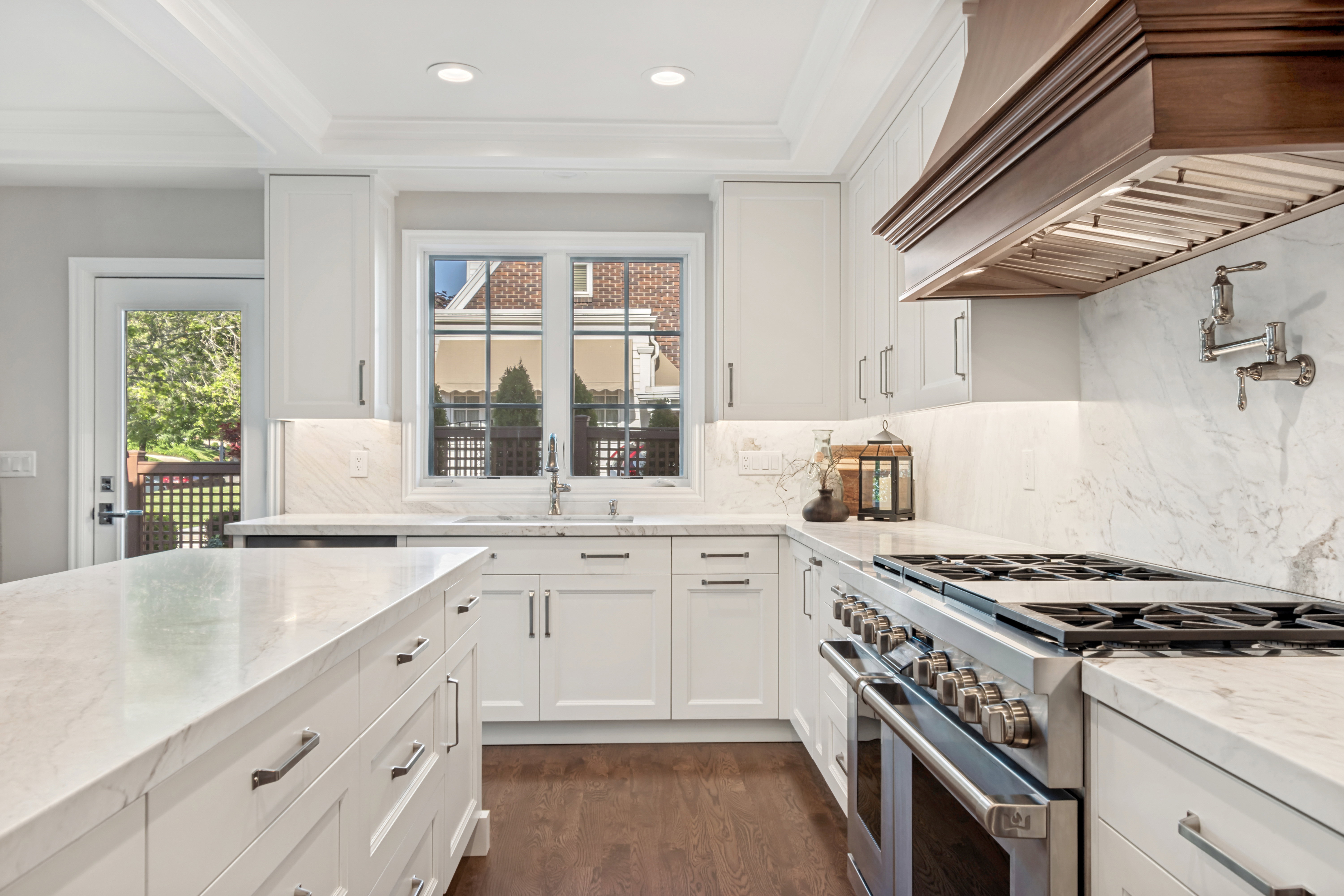 Gourmet kitchen featuring a large central island, white cabinetry with dark hardware, a professional-grade stainless steel range with a pot filler, a wood-finish range hood, and white stone countertops with matching backsplash