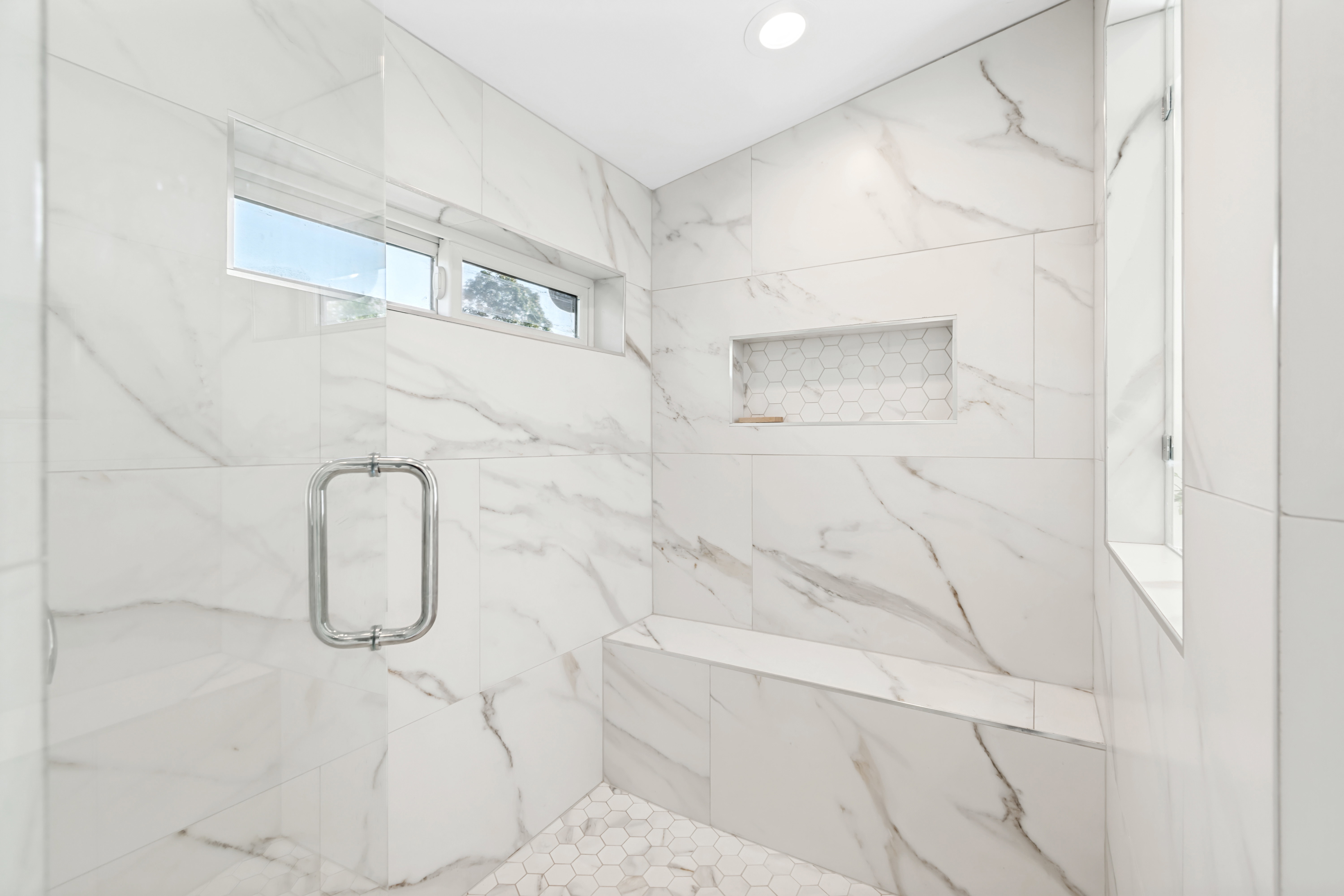Spacious walk-in shower featuring large format white tile with grey veining, a built-in bench, a tiled shower niche with a hexagonal accent, a transom window, and a clear glass shower door with chrome hardware
