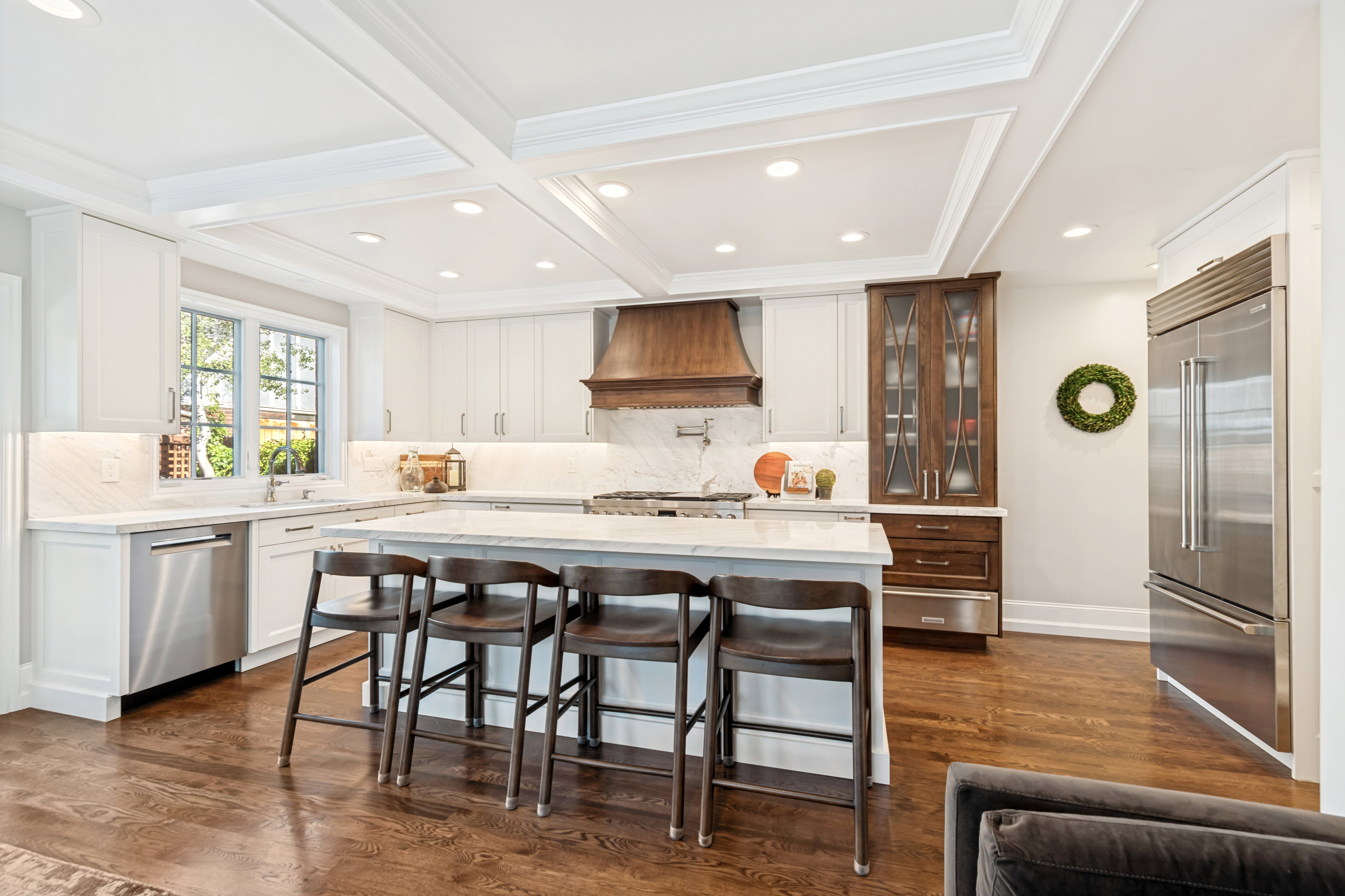 Gourmet kitchen featuring a substantial island with seating, a high-end stainless steel refrigerator, white cabinetry, a wood-finish range hood, and coffered ceilings