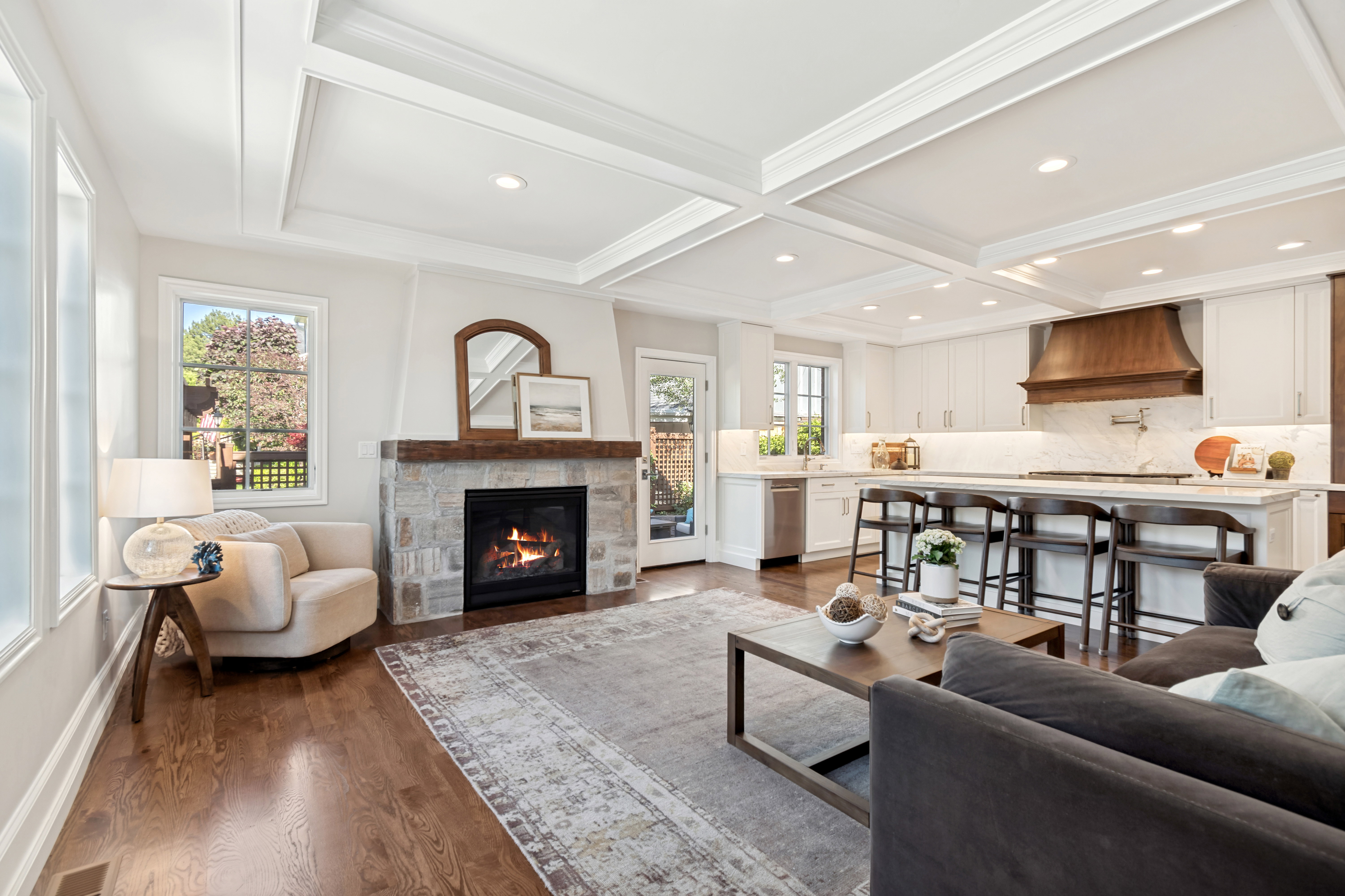 Spacious open-concept living area featuring rich wood-finish flooring, a stone fireplace with a substantial wood mantel, and crisp white coffered ceilings