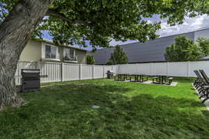 Expansive green lawn area featuring a white privacy fence, mature shade tree, multiple picnic tables on concrete pads, and a built-in barbecue grill