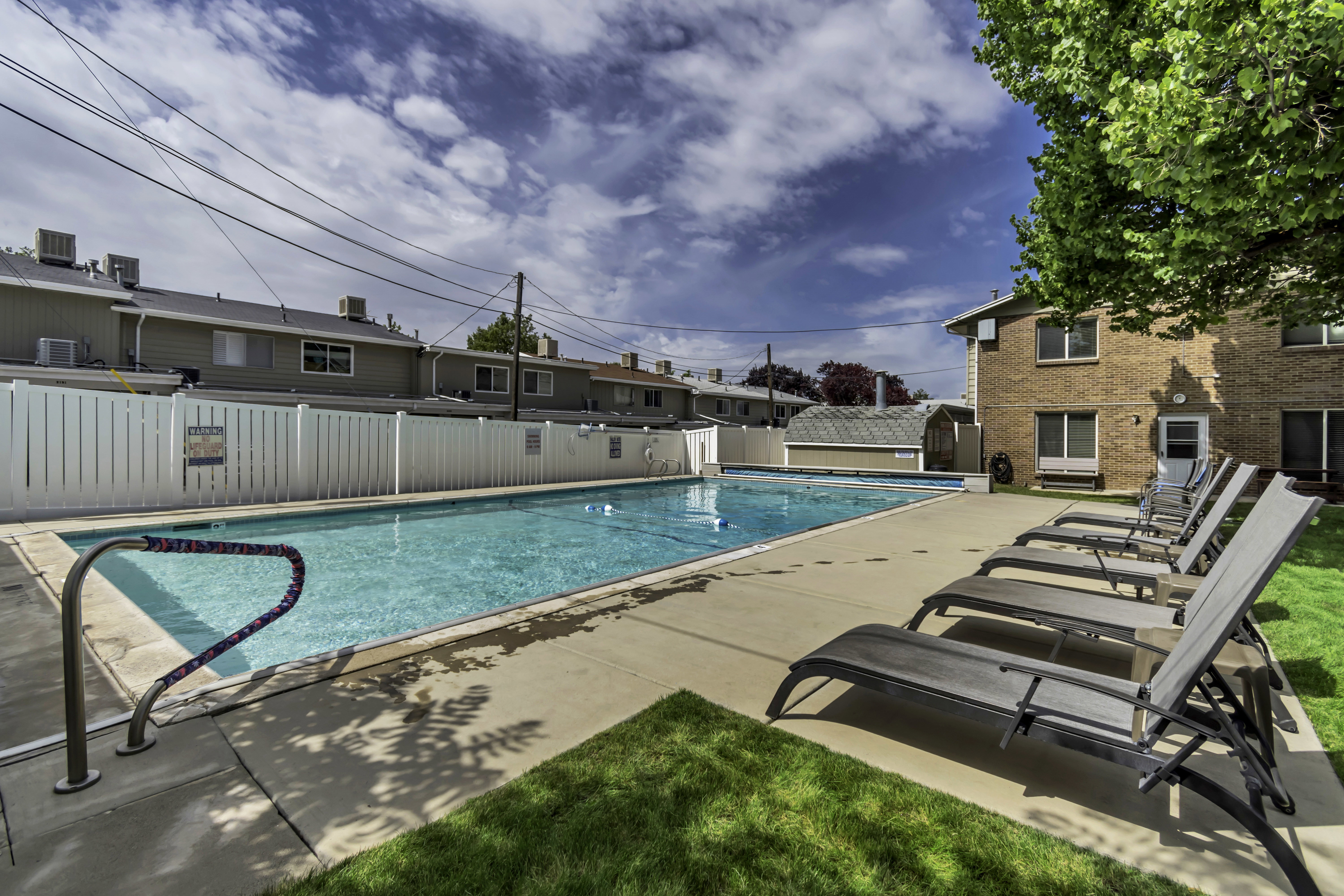 Expansive swimming pool area featuring a concrete deck, perimeter fencing, and a brick-exterior building