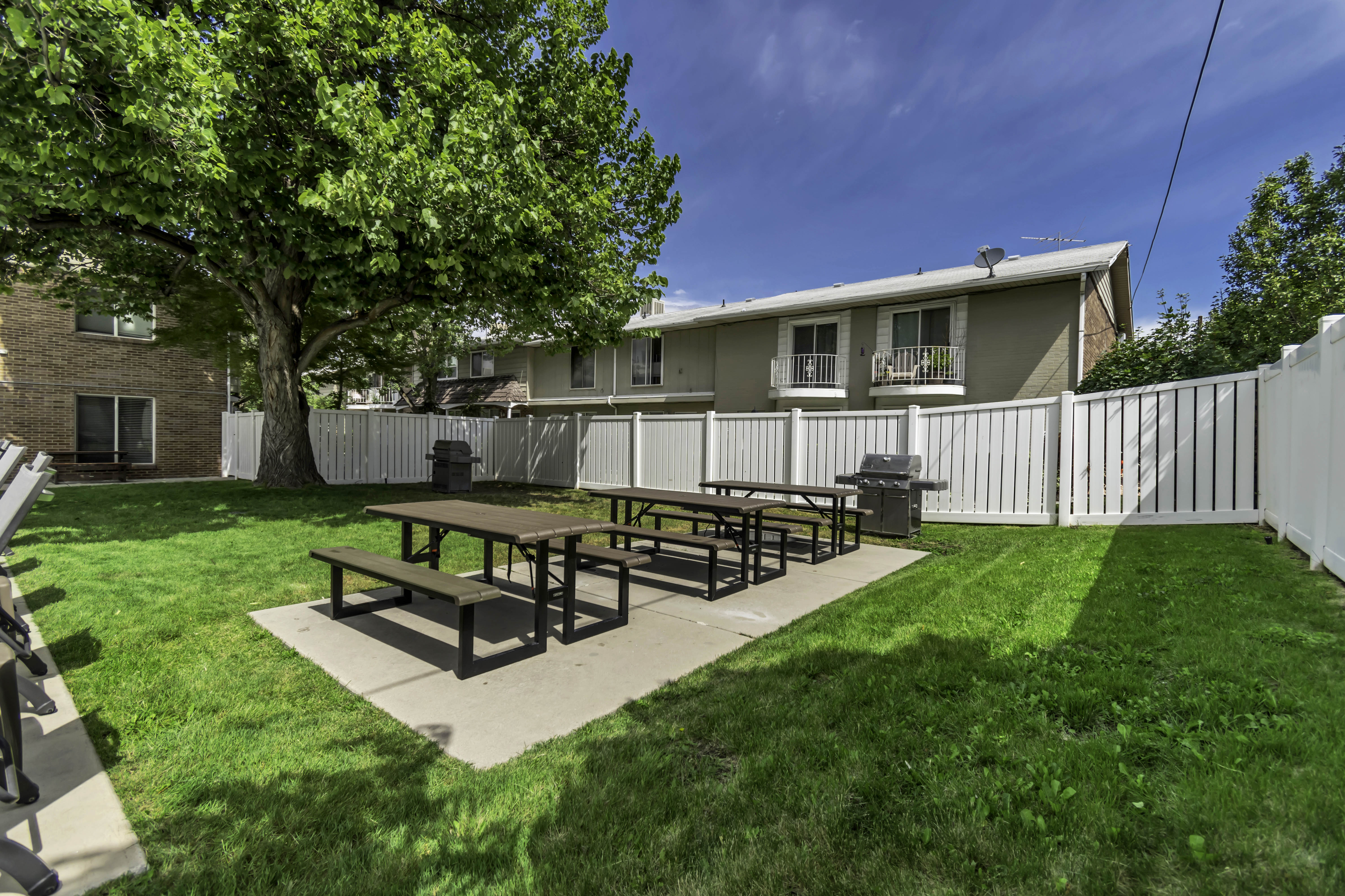 Shared outdoor space featuring a lush lawn, concrete patio areas, two picnic table sets, and a white privacy fence