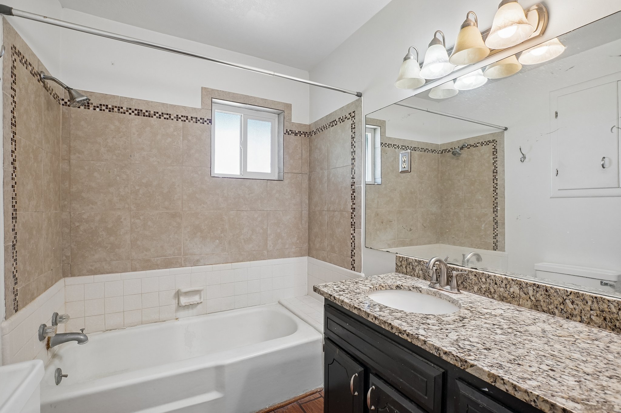 Bathroom featuring a built-in tub with tile surround, a window over the tub, a vanity with a granite-style countertop, an undermount sink, and an expansive mirror