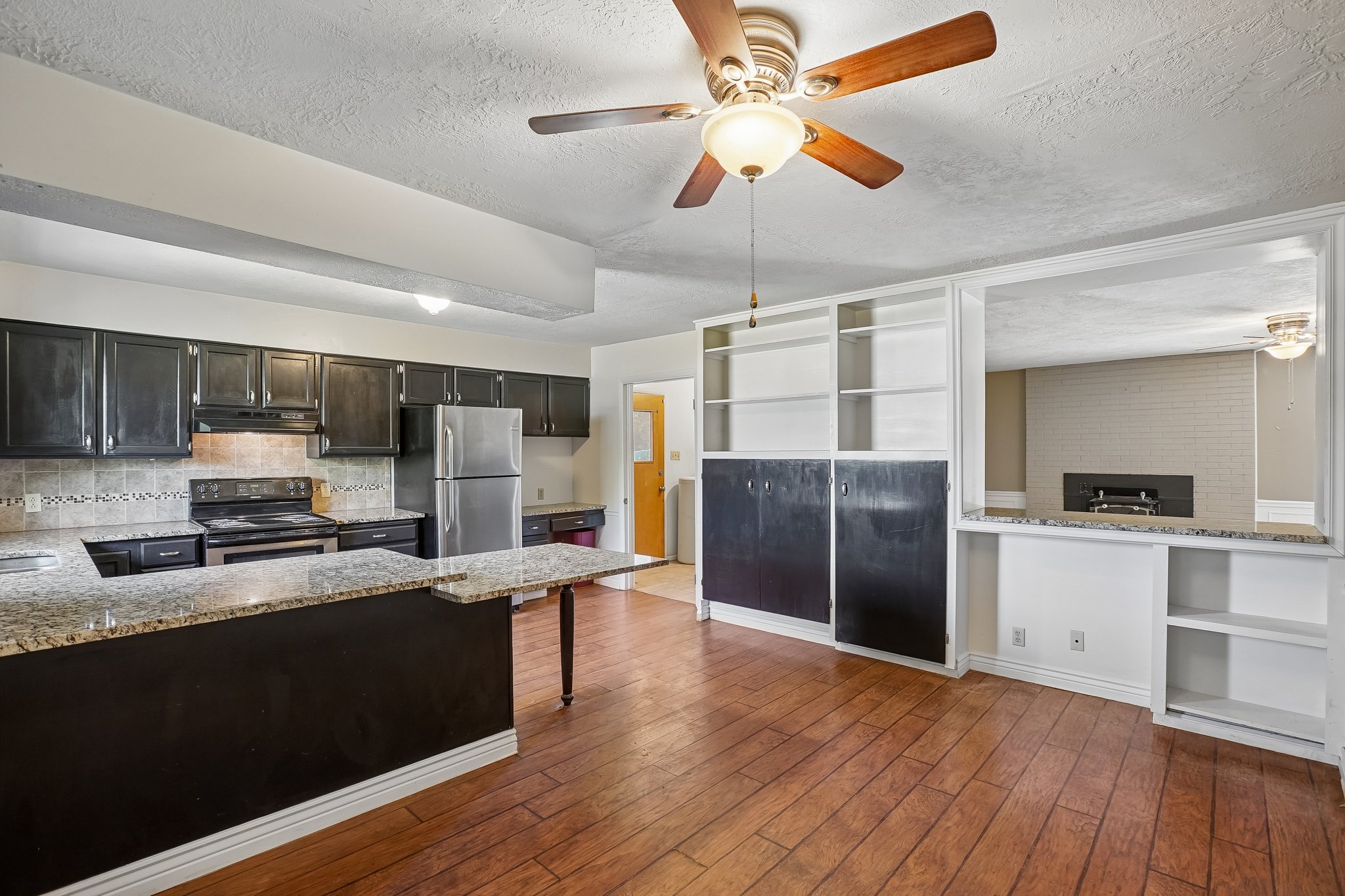 Open-concept living area with wood-finish flooring, a ceiling fan with light fixture, and a kitchen featuring dark cabinetry, stainless steel appliances, and light-toned countertops