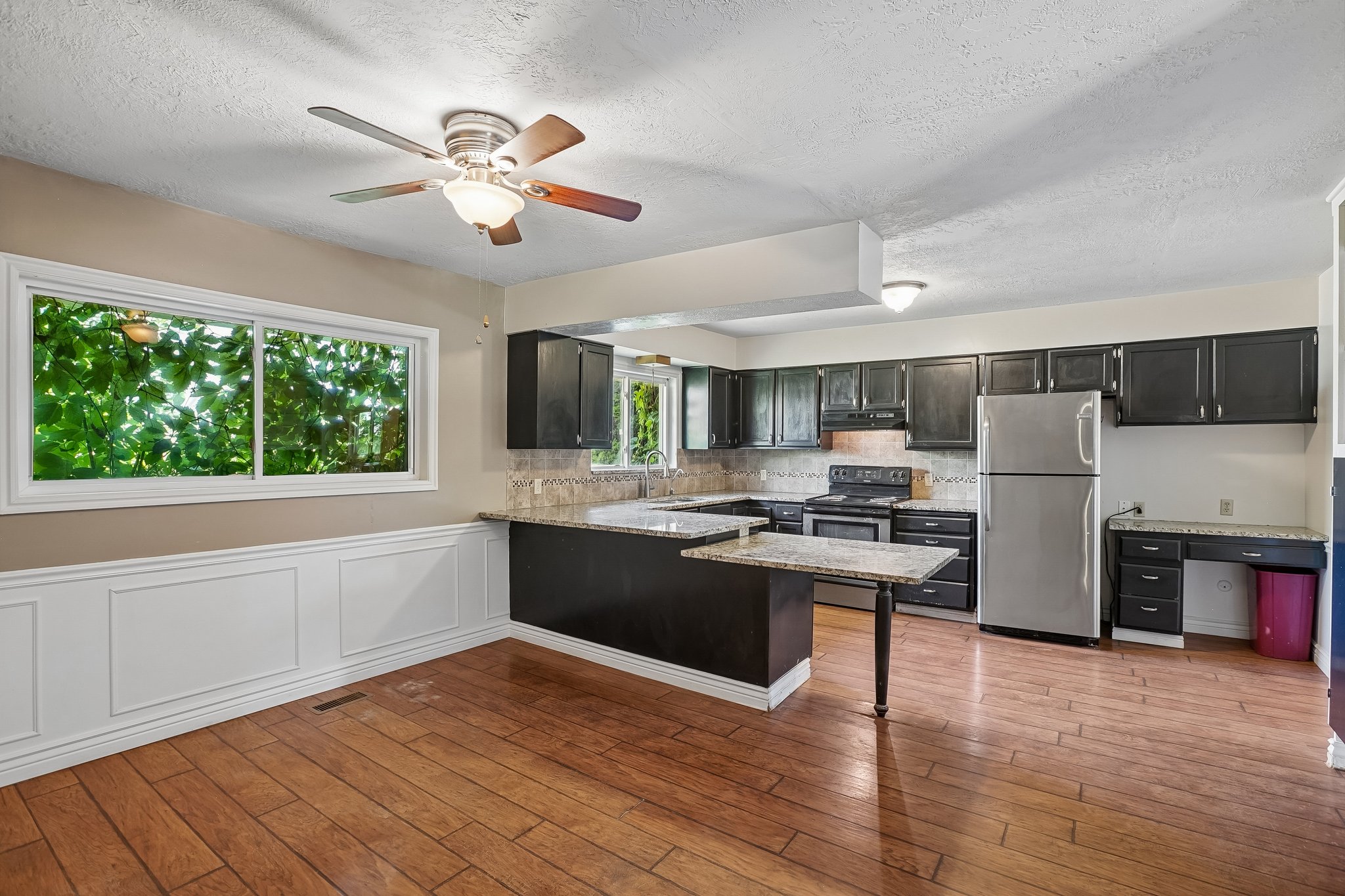 Open-concept kitchen and dining area featuring wood-finish flooring, a ceiling fan, and a large window overlooking lush greenery
