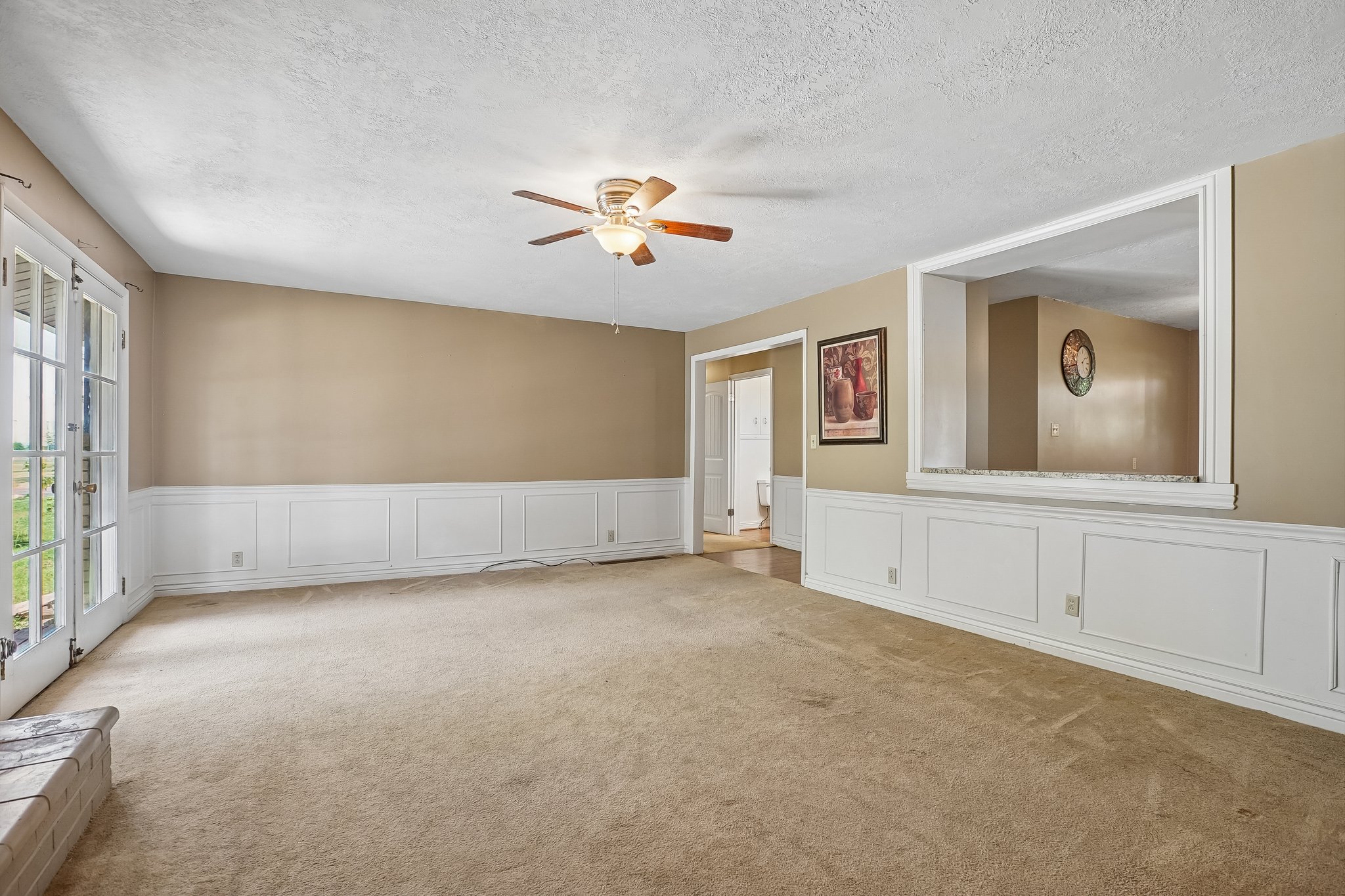 Spacious living area featuring neutral carpeting, a ceiling fan with light fixture, and wainscoting with a chair rail