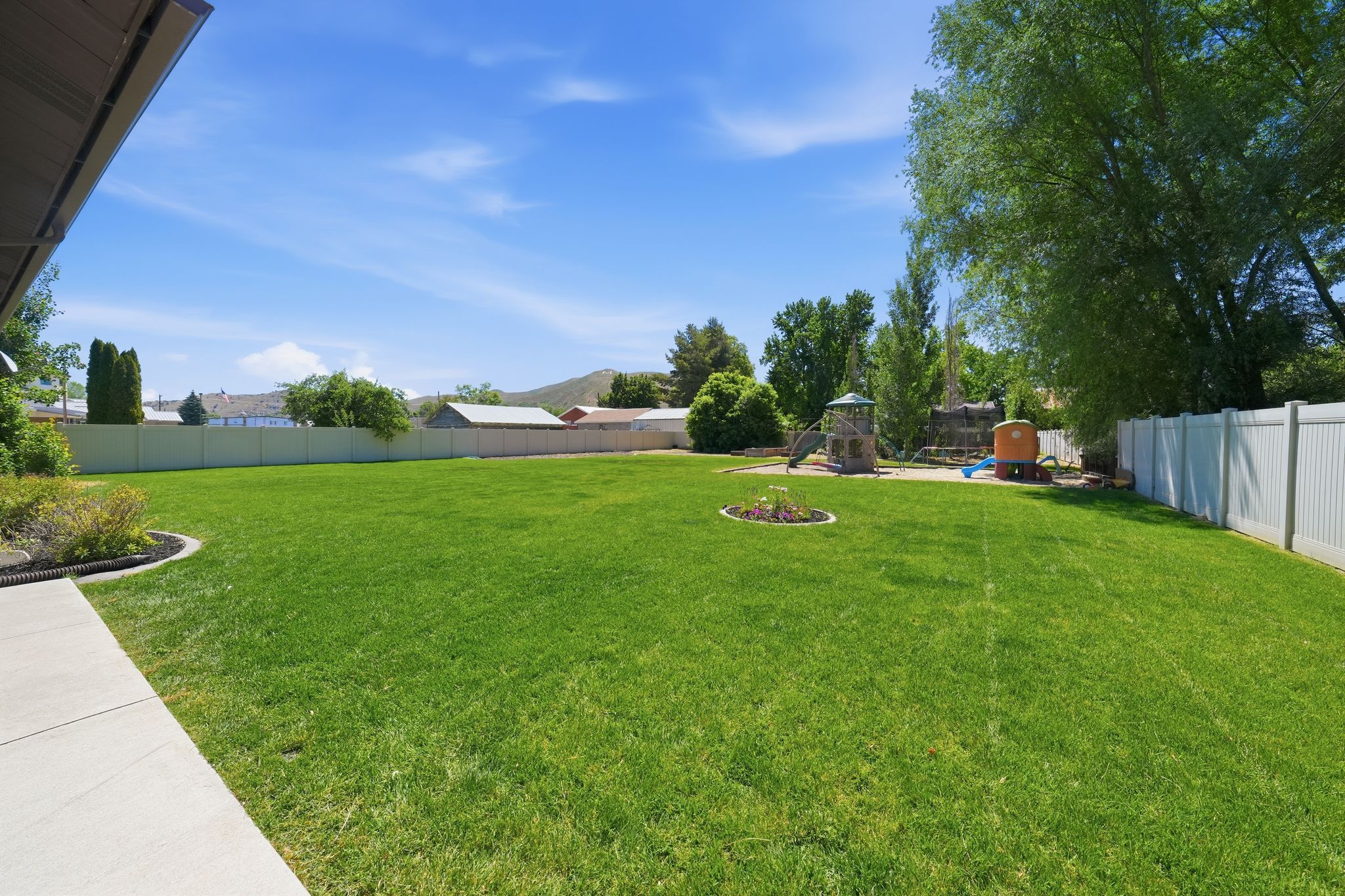 Expansive backyard featuring a lush lawn, white vinyl fencing, mature trees, and a paved patio area