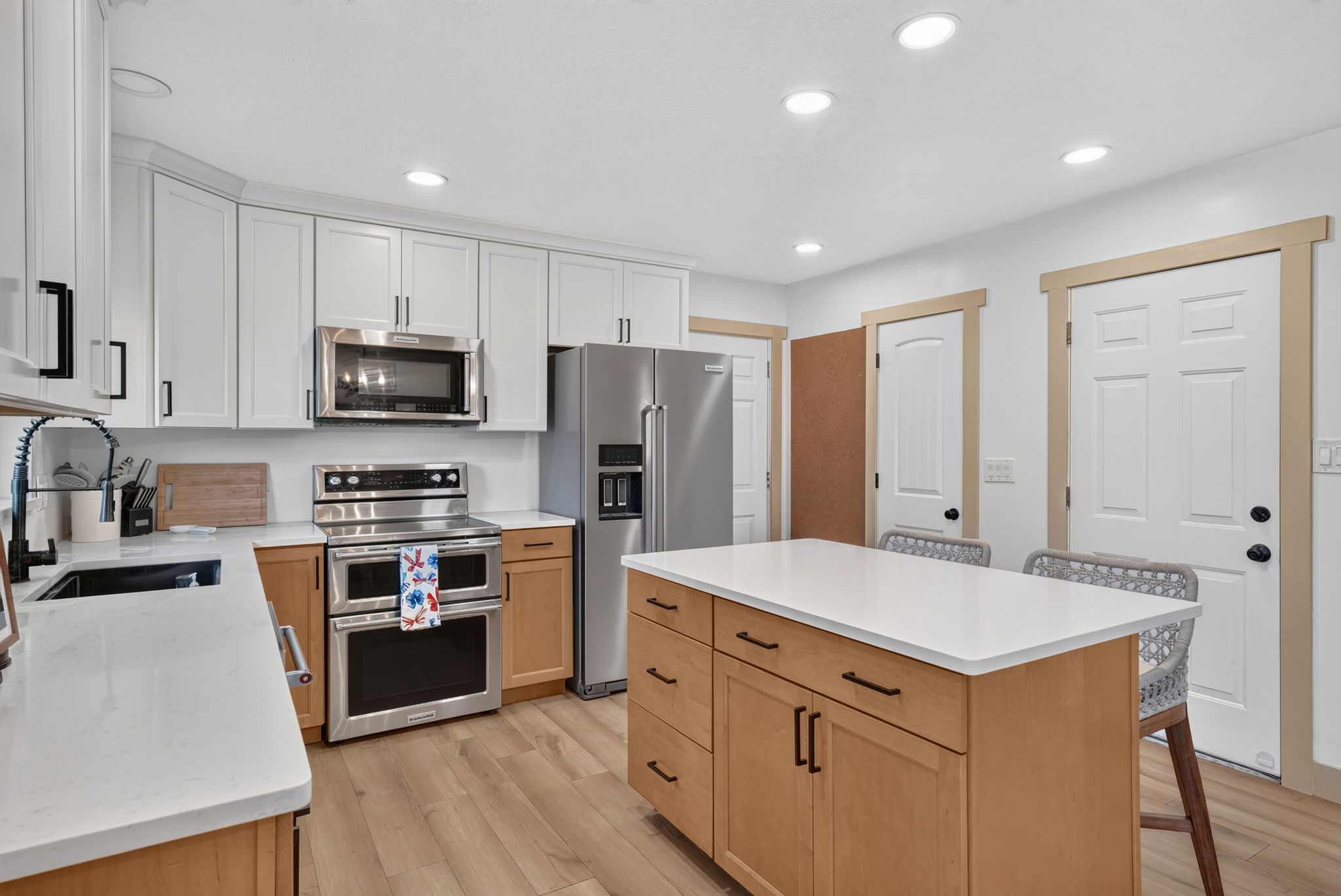 Modern kitchen featuring a central island with white countertop, contrasting two-tone cabinetry, stainless steel appliances, wood-finish flooring, and recessed lighting